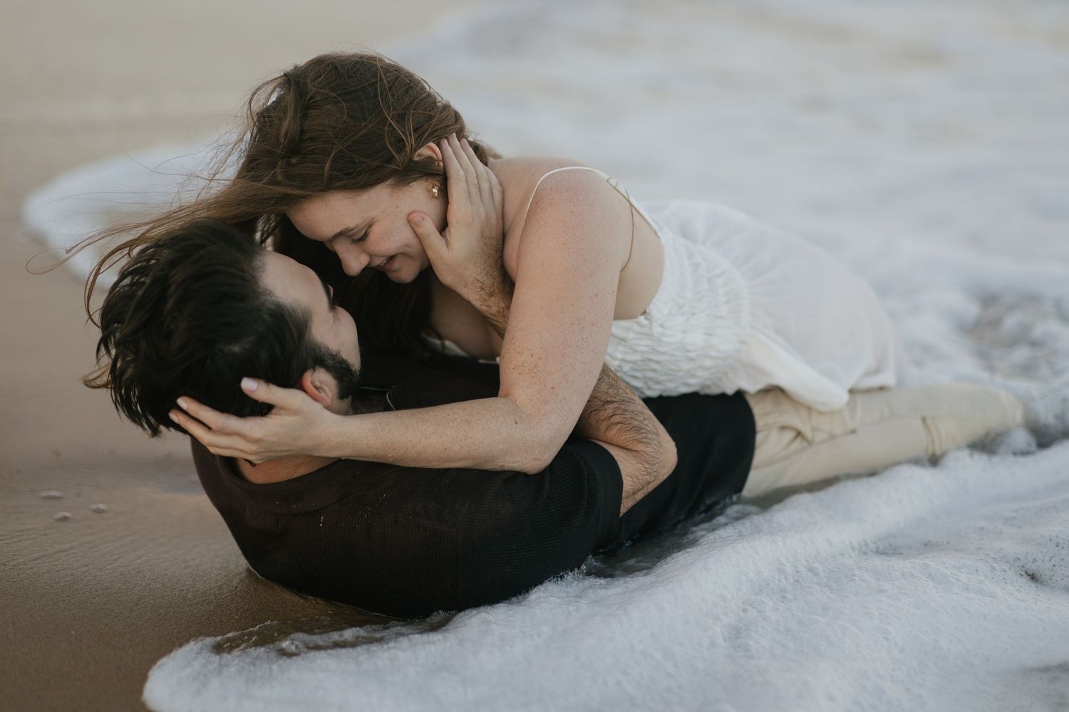 Couple embracing romantically on a sandy beach with ocean waves washing around them.