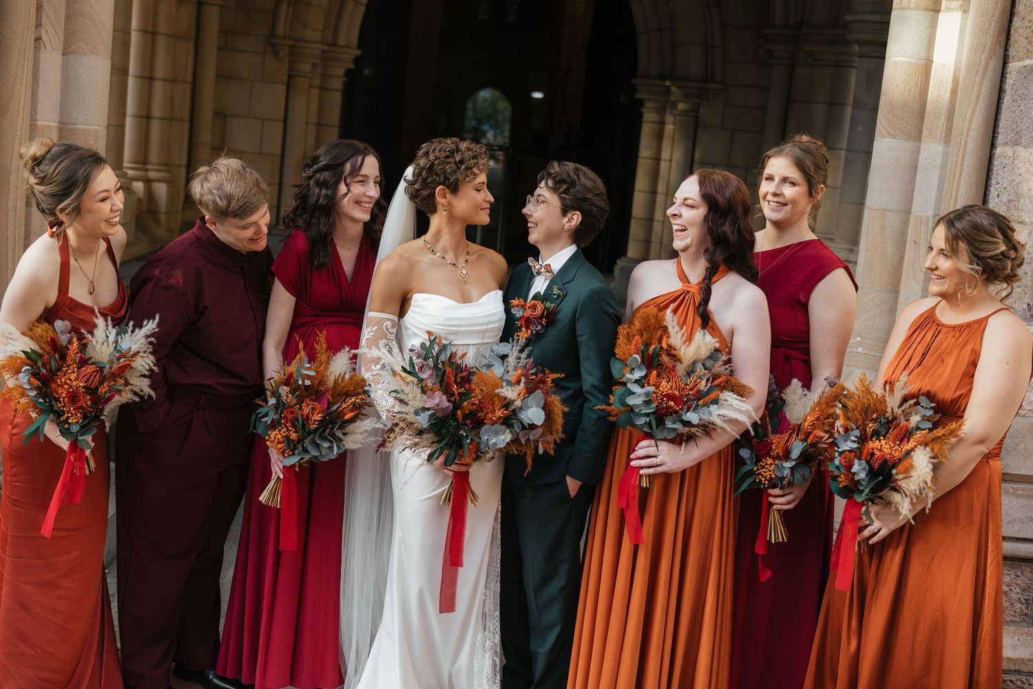 Bride and bridesmaids in red and orange dresses holding autumn bouquets outside a stone church.