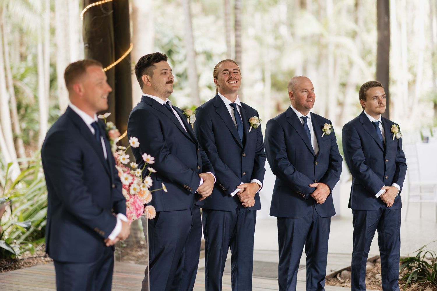 Five groomsmen in navy suits with floral boutonnieres laughing and smiling during an outdoor wedding ceremony.