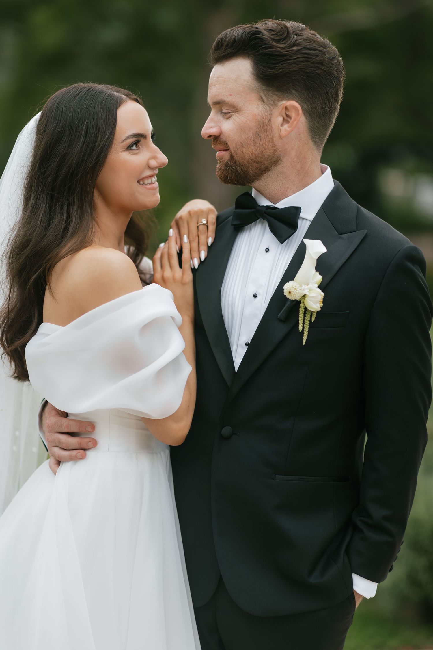 Bride in off-shoulder white gown and groom in black tuxedo share a romantic gaze outdoors on their wedding day.