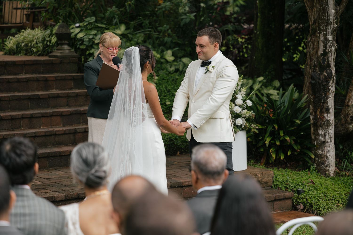 Bride and groom holding hands during outdoor wedding ceremony with officiant and seated guests in lush garden setting.