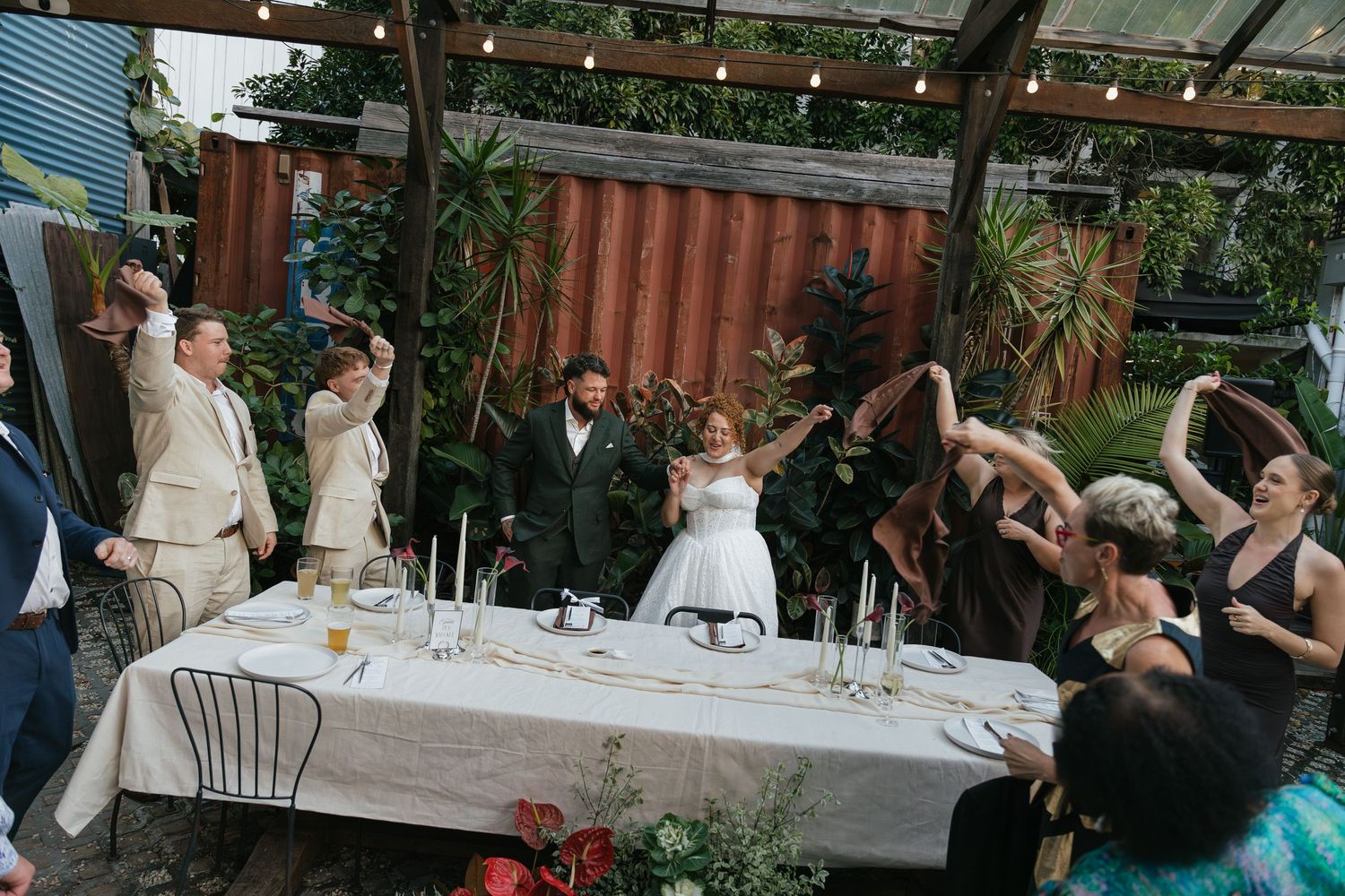 Wedding party raises glasses in a toast at an outdoor reception table adorned with candles and greenery.