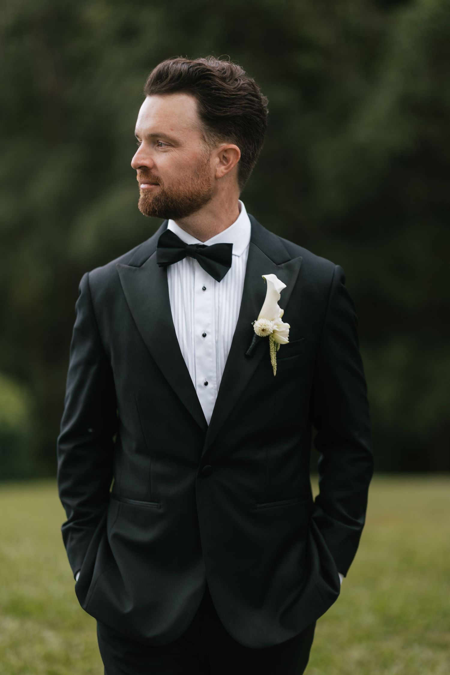 A dapper groom in a black tuxedo with bow tie and white boutonniere stands outdoors with hands in pockets.
