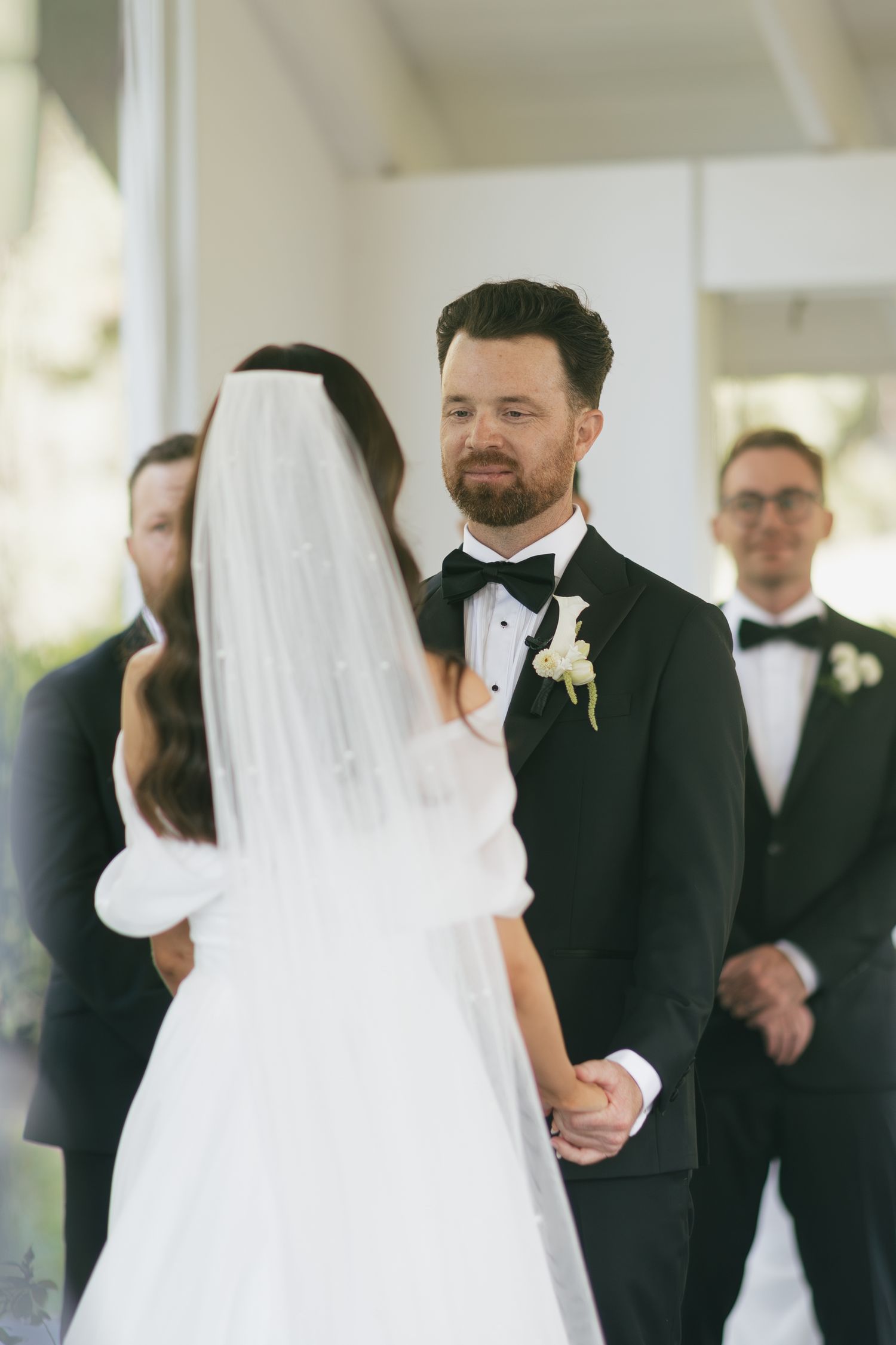 Groom in black tuxedo smiling at bride in white gown and veil during outdoor wedding ceremony.