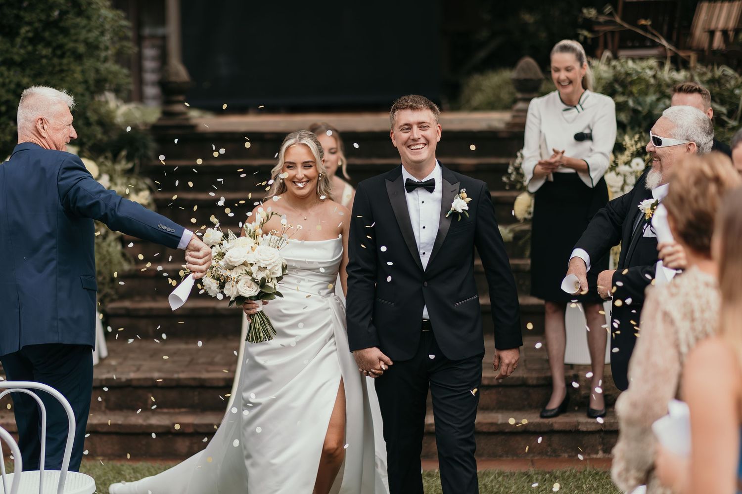 Bride and groom walk down the aisle as guests toss confetti at an elegant outdoor wedding ceremony.