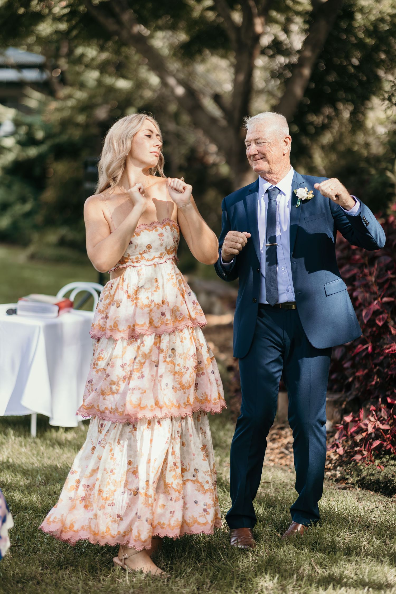 A young woman in a floral dress dances with an older man in a navy suit at an outdoor wedding reception.