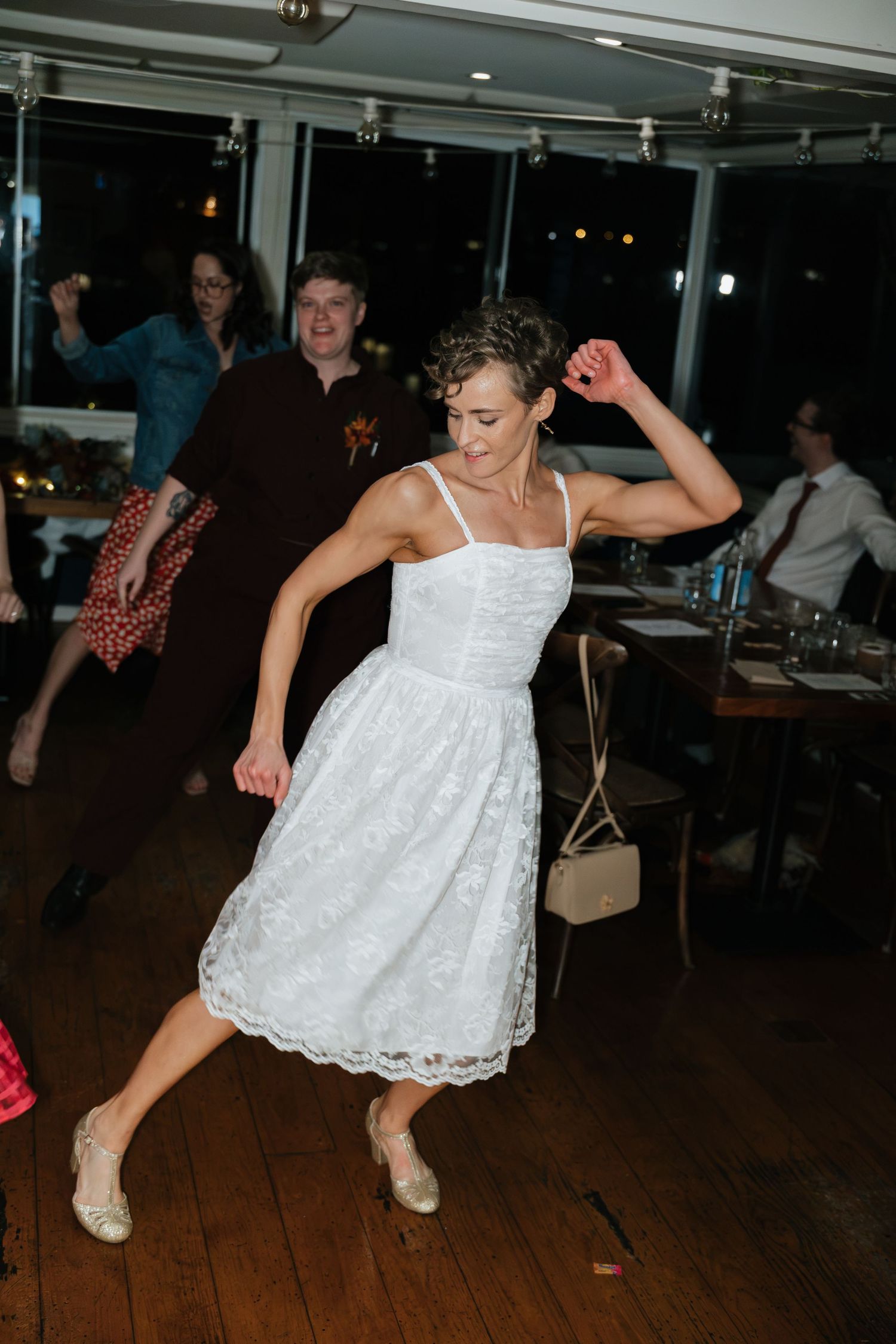 Bride in white lace dress dancing joyfully at wedding reception with guests celebrating in background.