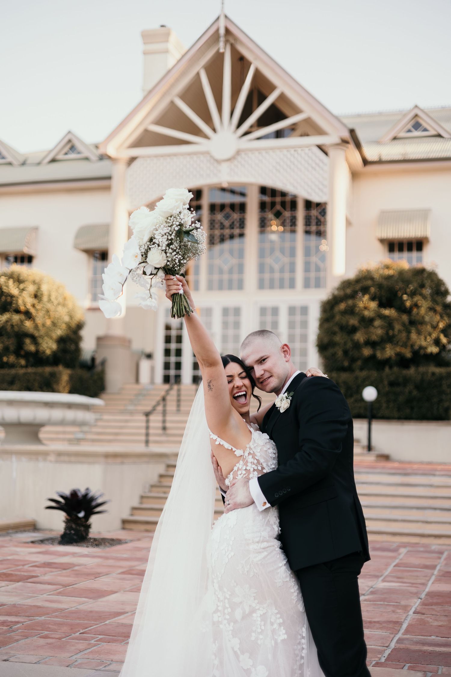 Newlywed couple dances and twirls together on a tiled terrace with elegant architecture in the background at sunset.