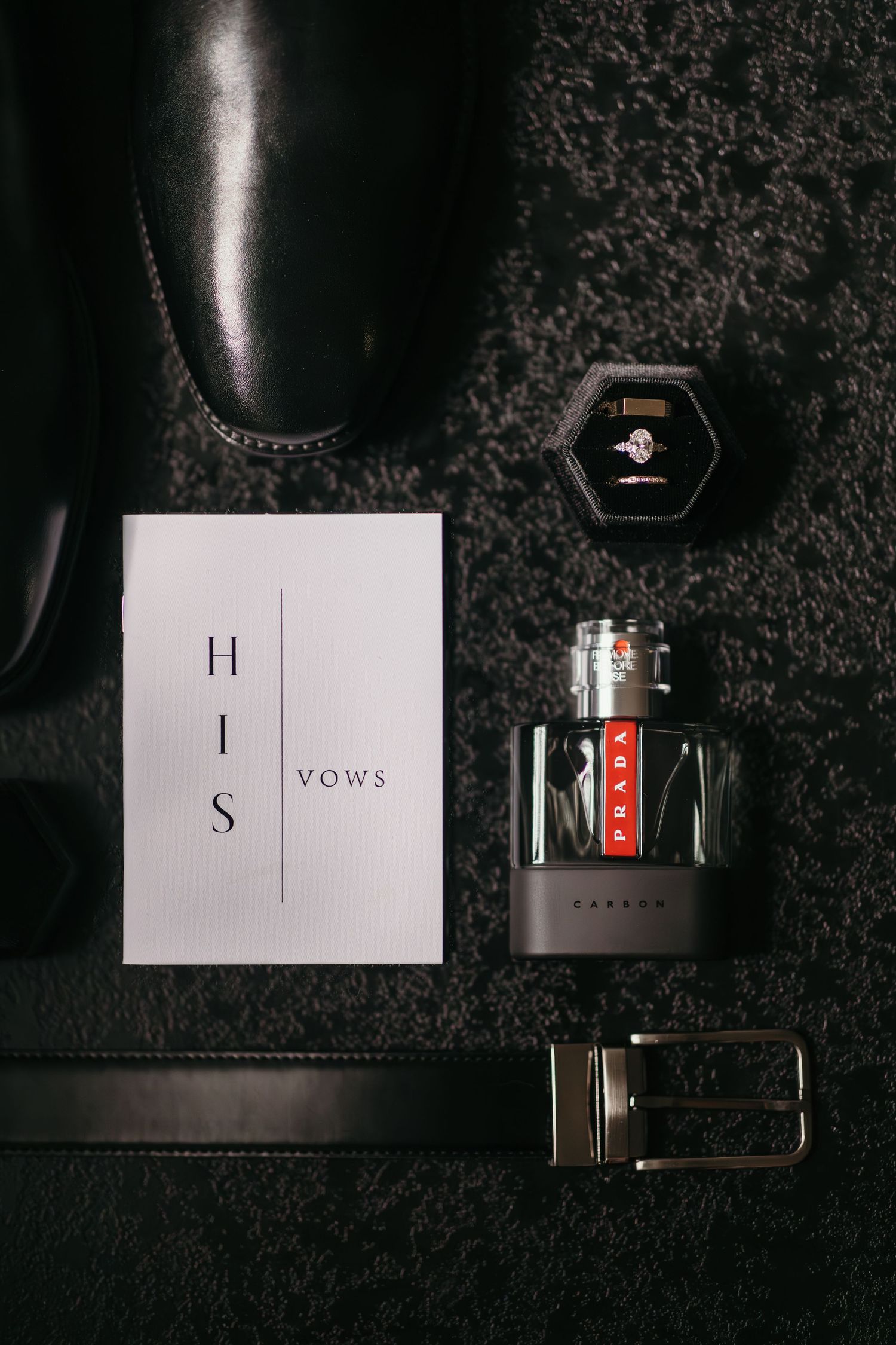 Groom wedding flat lay with black dress shoes, HIS vows card, Prada cologne, leather belt, and diamond ring in box.