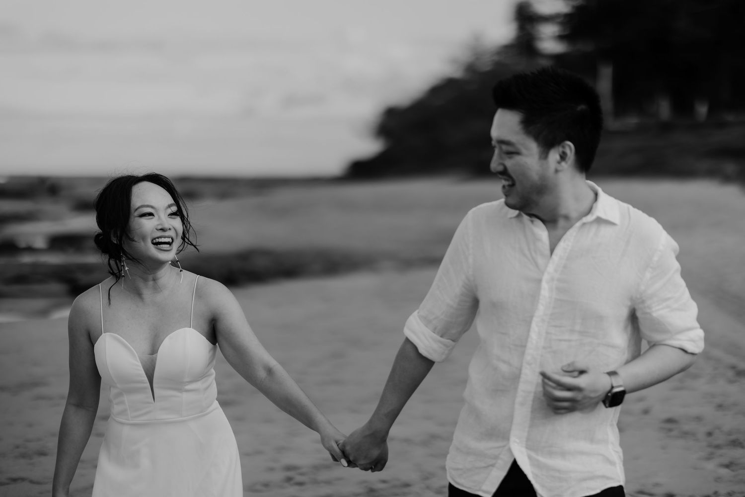 A joyful couple holds hands on a beach, the woman in a strapless dress laughing as they walk together.