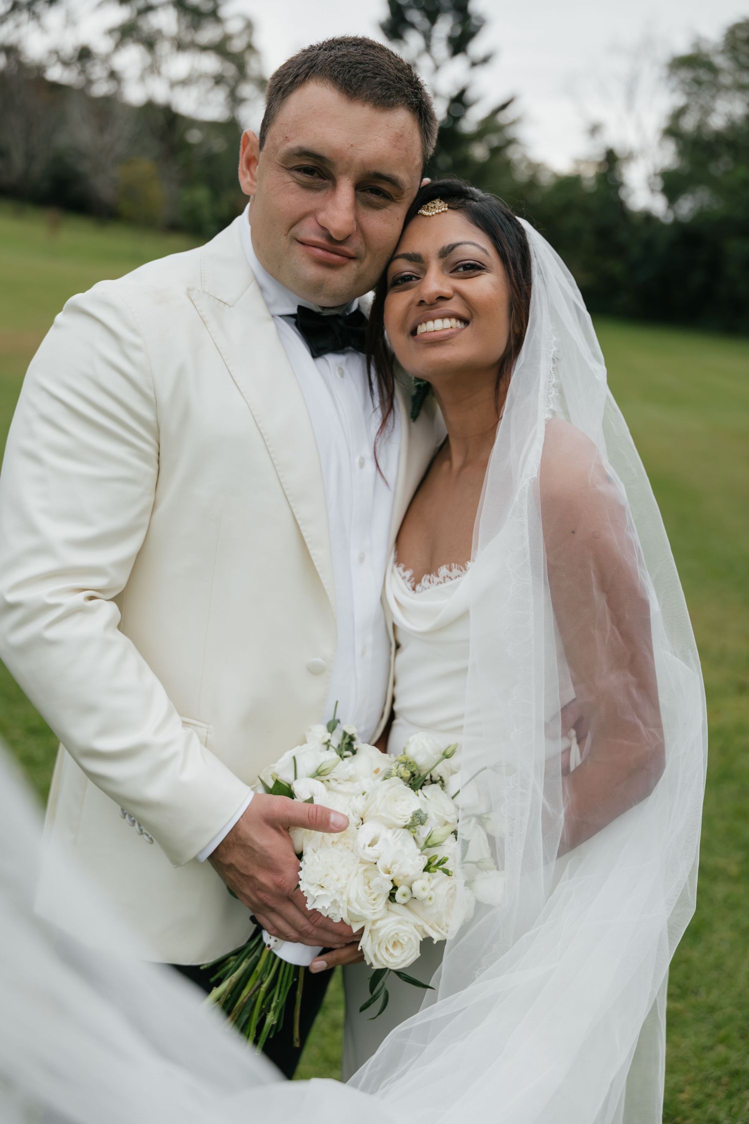 A smiling bride and groom in white wedding attire pose outdoors, she holds a white rose bouquet with veil flowing.