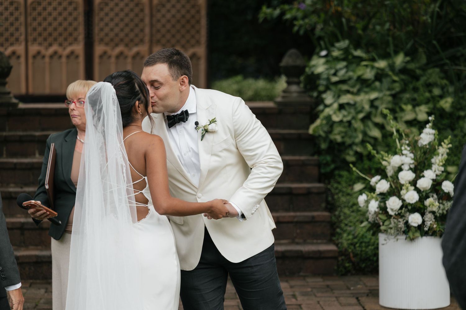 Groom in white tuxedo jacket kissing bride in white gown and veil during outdoor wedding ceremony.