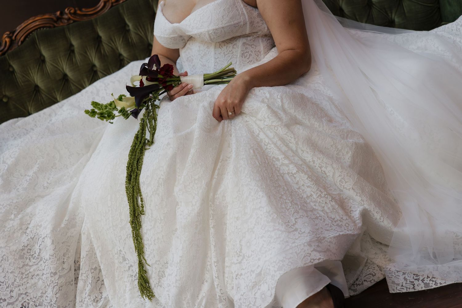 Bride in white lace wedding gown holding elegant bouquet with trailing greenery, seated on green velvet sofa.
