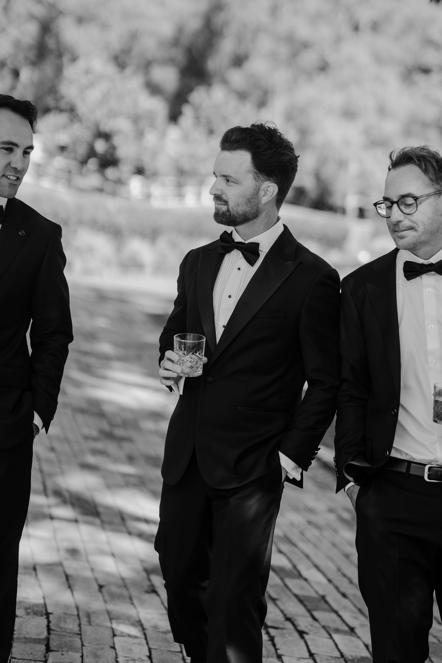 Three groomsmen in black tuxedos with bow ties walk outdoors, one holding a glass, in a black and white photo.