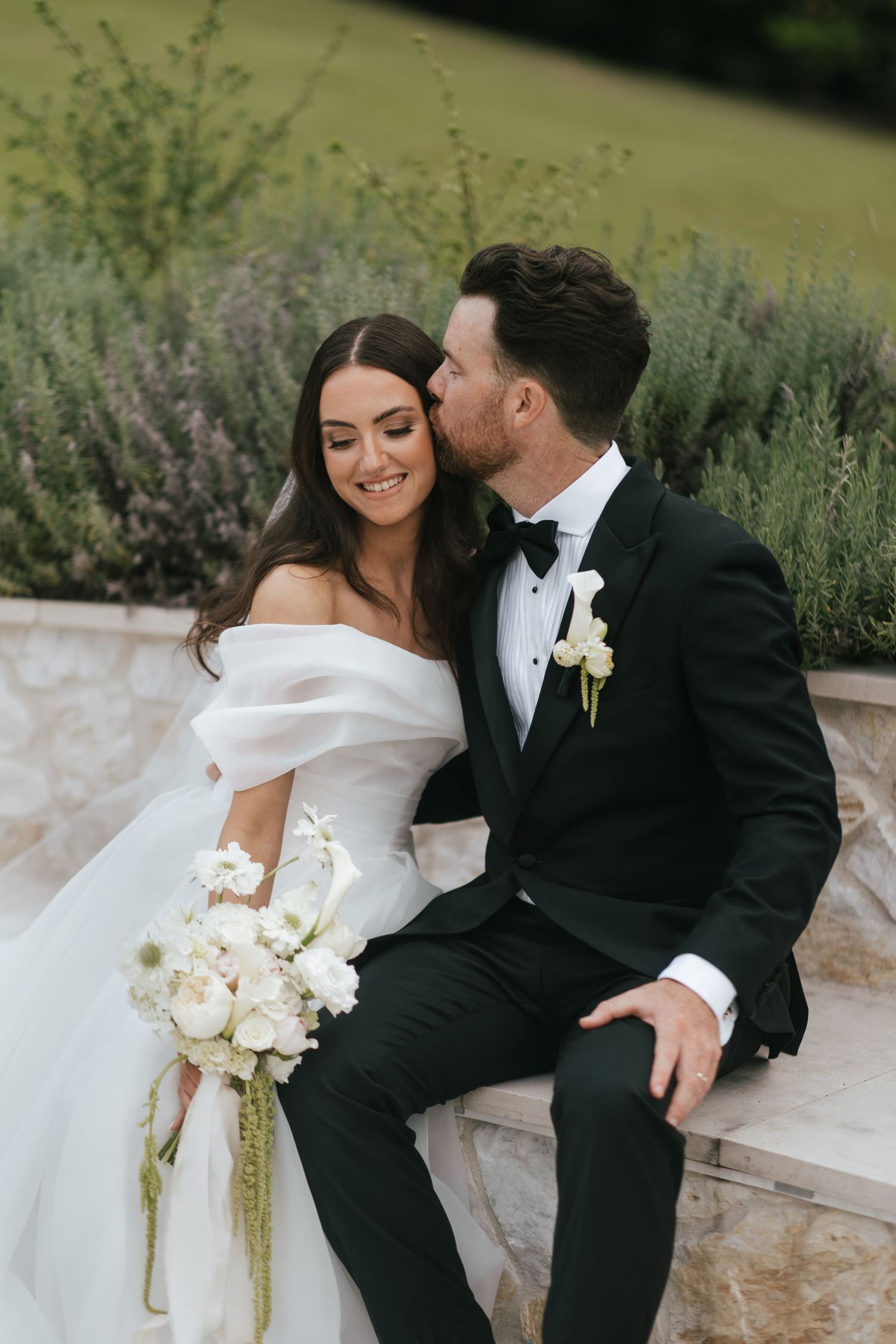 Bride in off-shoulder gown holds white bouquet as groom in black tuxedo kisses her cheek outdoors.