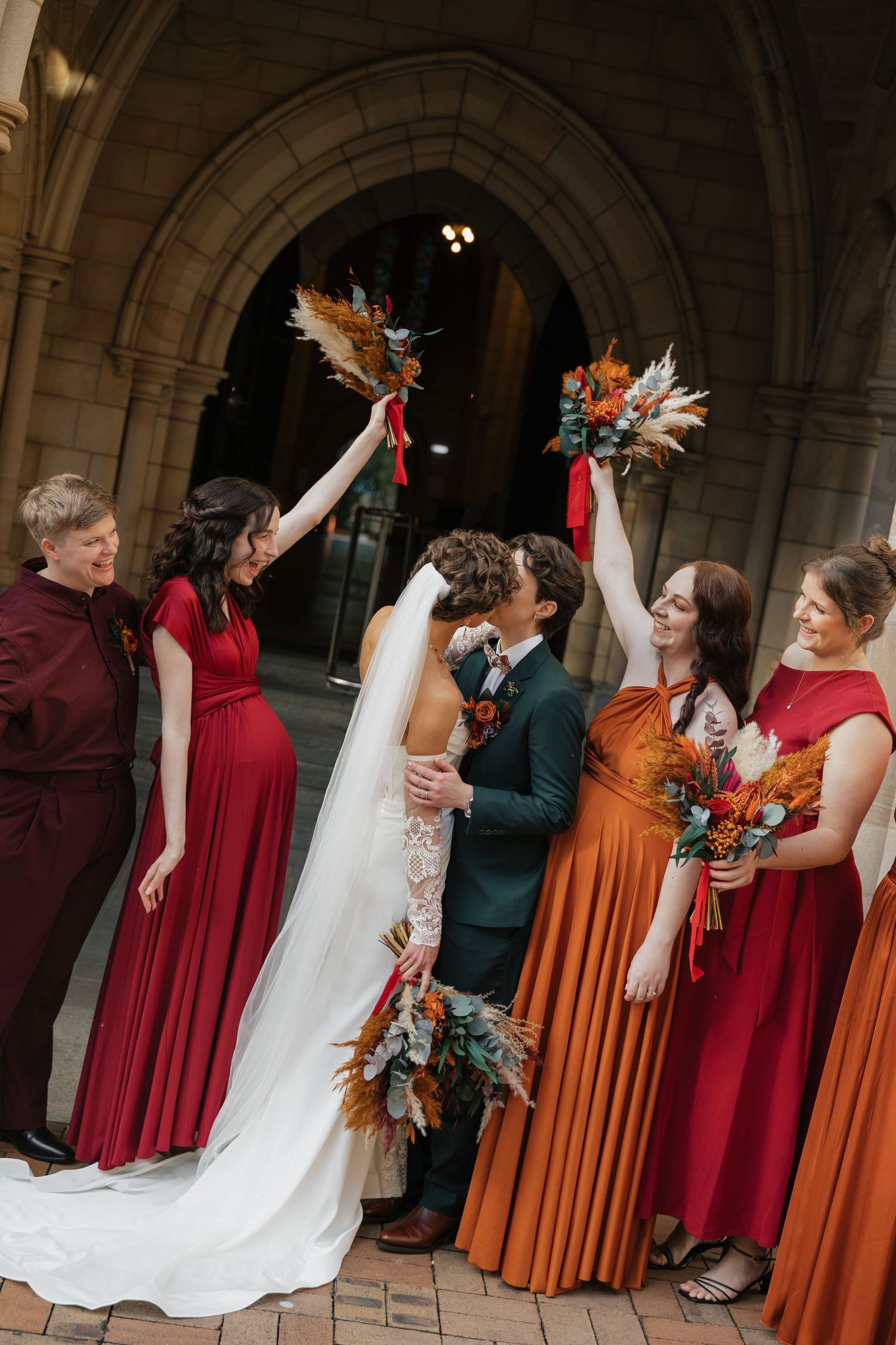 Wedding party celebrates with raised bouquets as bride and groom kiss, bridesmaids in red and orange gowns outside stone arch.