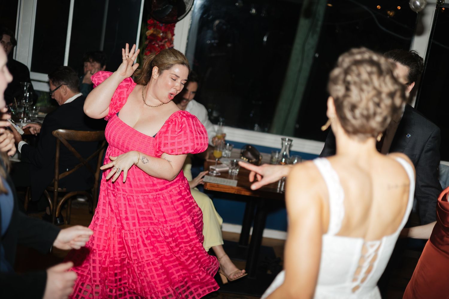 Woman in pink ruffled dress dancing joyfully at a wedding reception with bride in white dress.