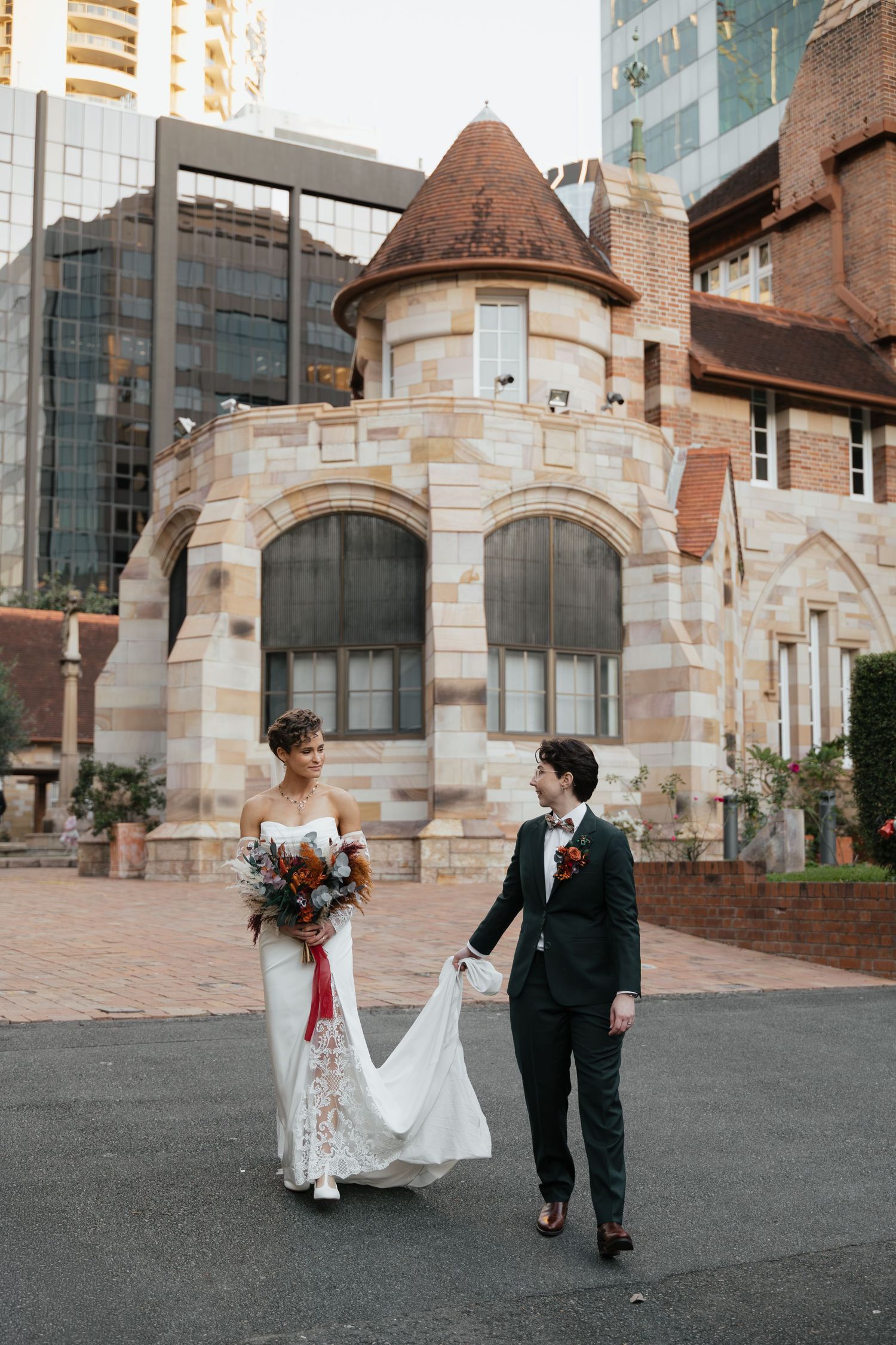 Bride and groom walking in front of historic stone building with modern skyscrapers, bride holding floral bouquet.