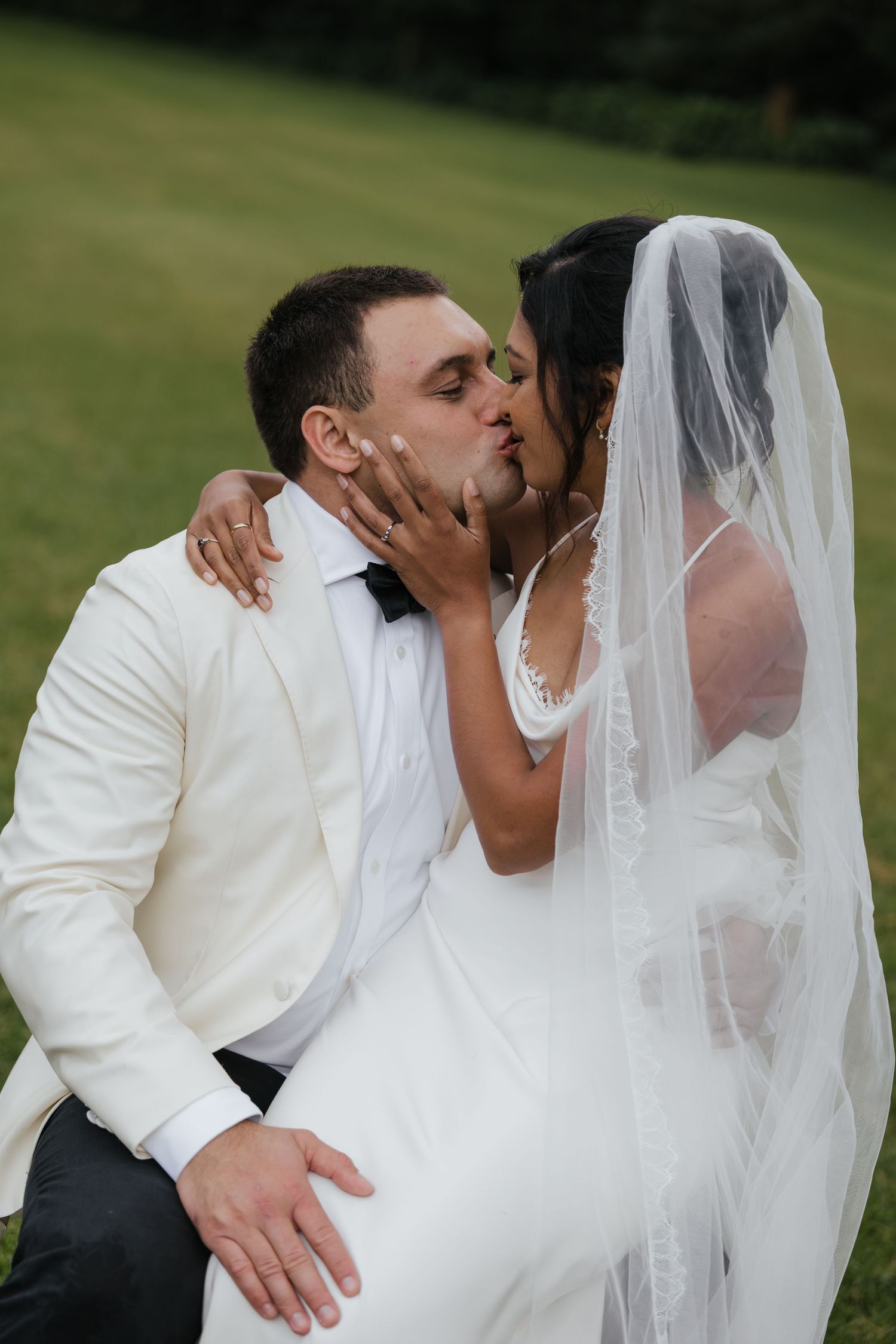 A bride and groom share a romantic kiss outdoors, with the bride wearing a flowing veil and white dress on their wedding day.