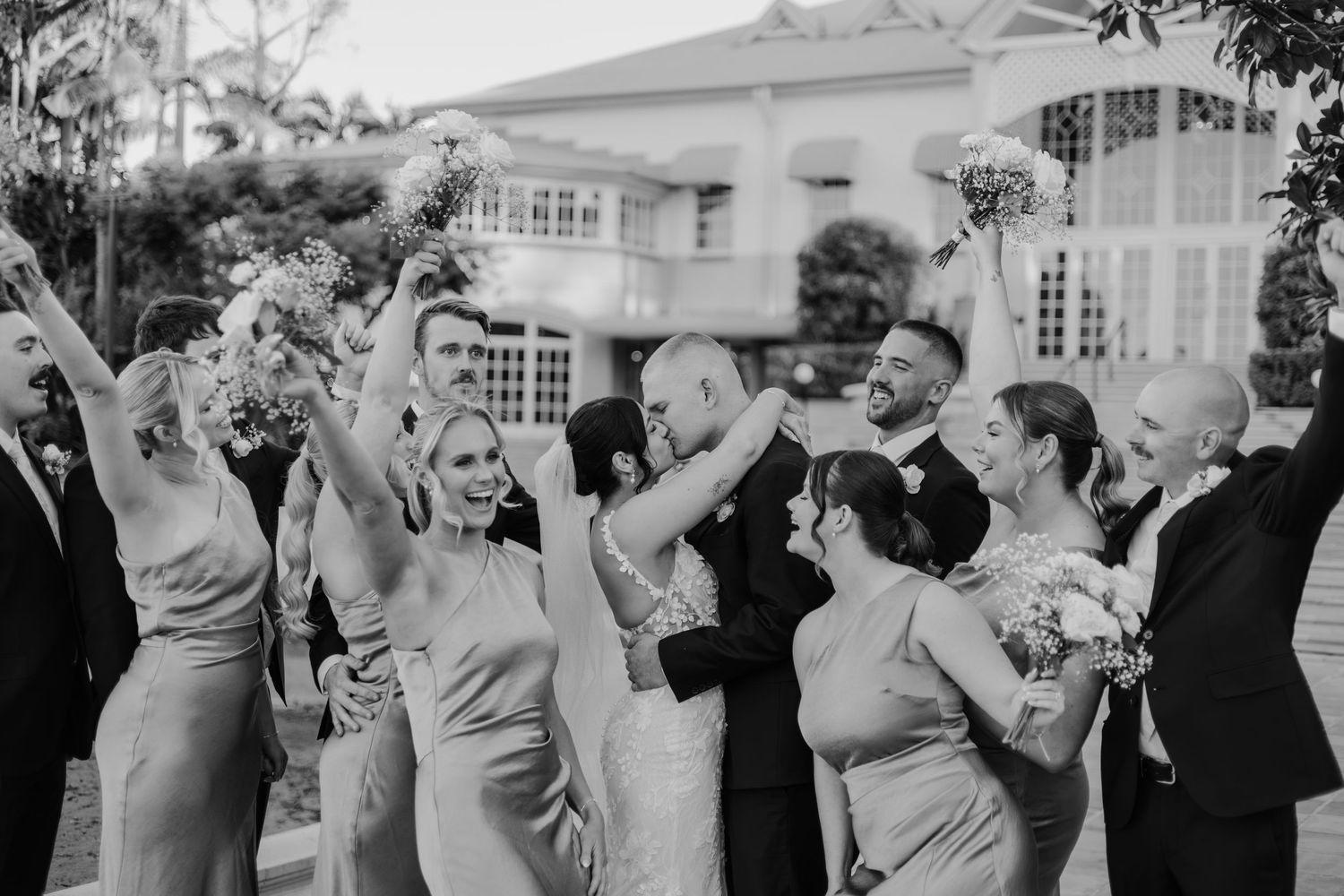 Black and white wedding party photo with bride and groom kissing surrounded by cheering bridesmaids and groomsmen.