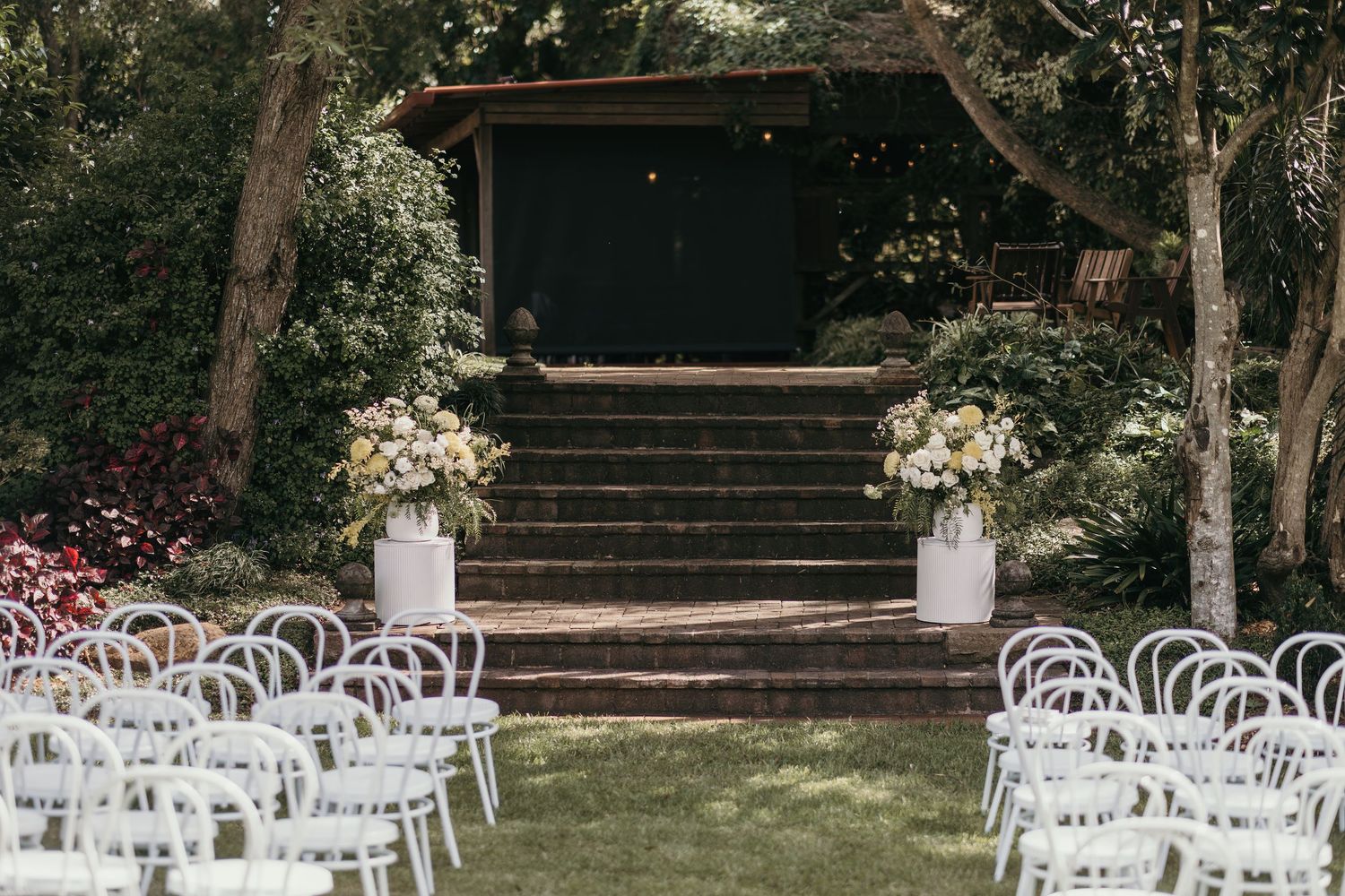 Elegant outdoor wedding ceremony setup with white chairs, floral arrangements, and rustic wooden steps surrounded by lush greenery.