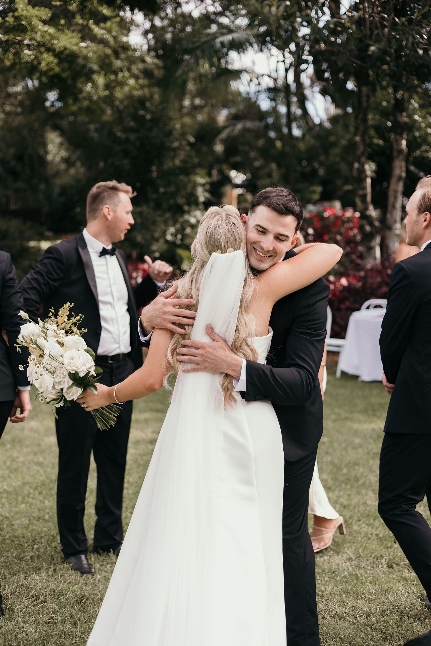 Bride in white gown hugs smiling groom in black tuxedo outdoors at elegant garden wedding ceremony.