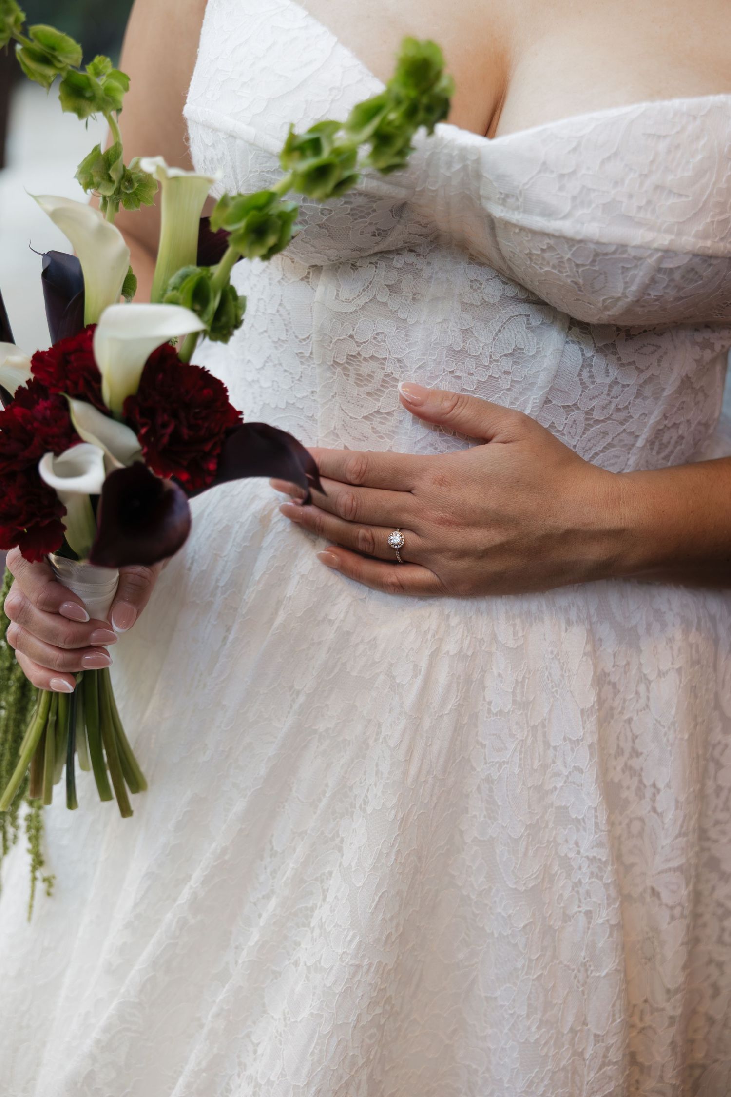 Pregnant bride in white lace dress holding red and white calla lily bouquet, with groom's hand on her baby bump.