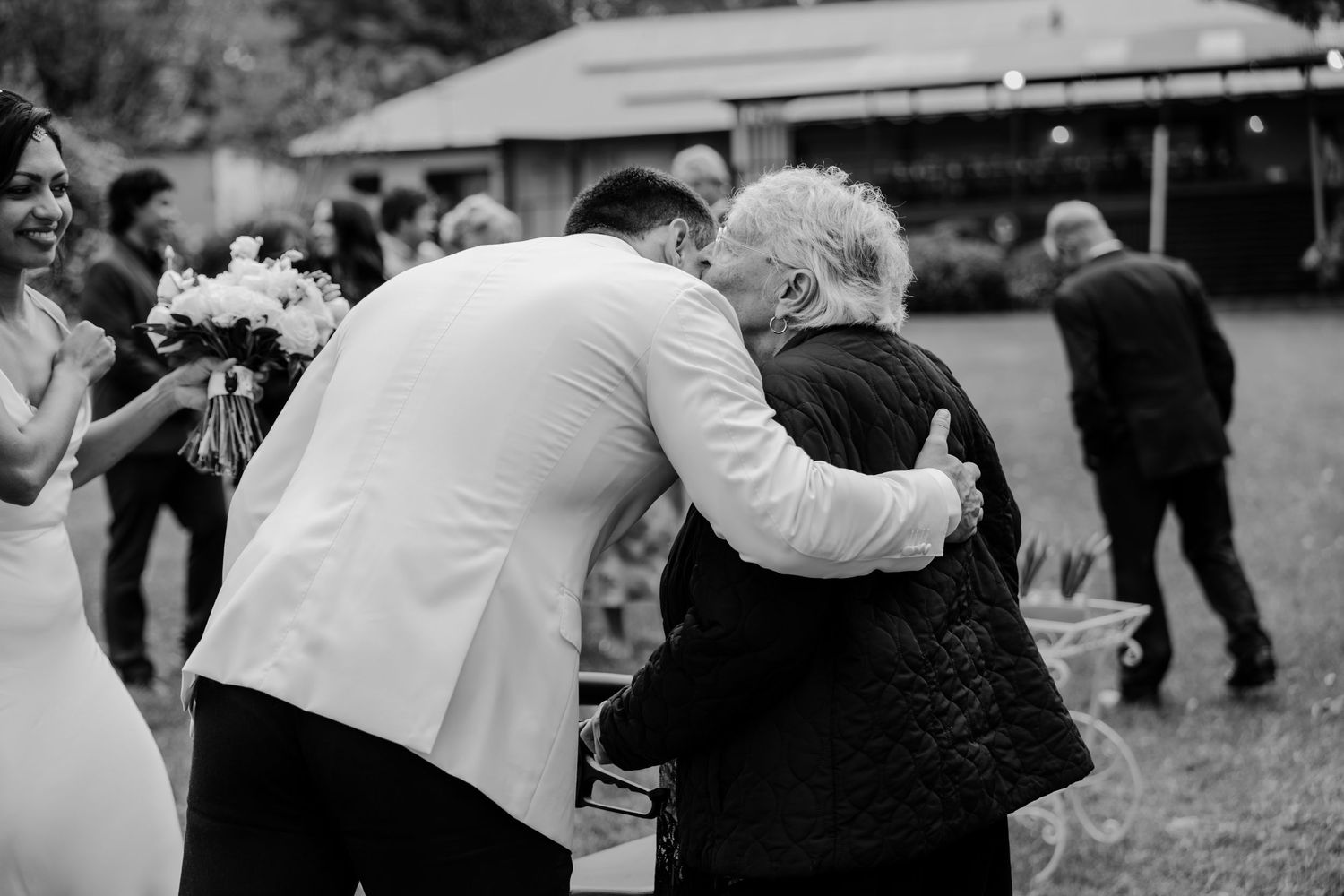 Groom in white jacket embracing elderly guest at outdoor wedding reception in black and white photo.