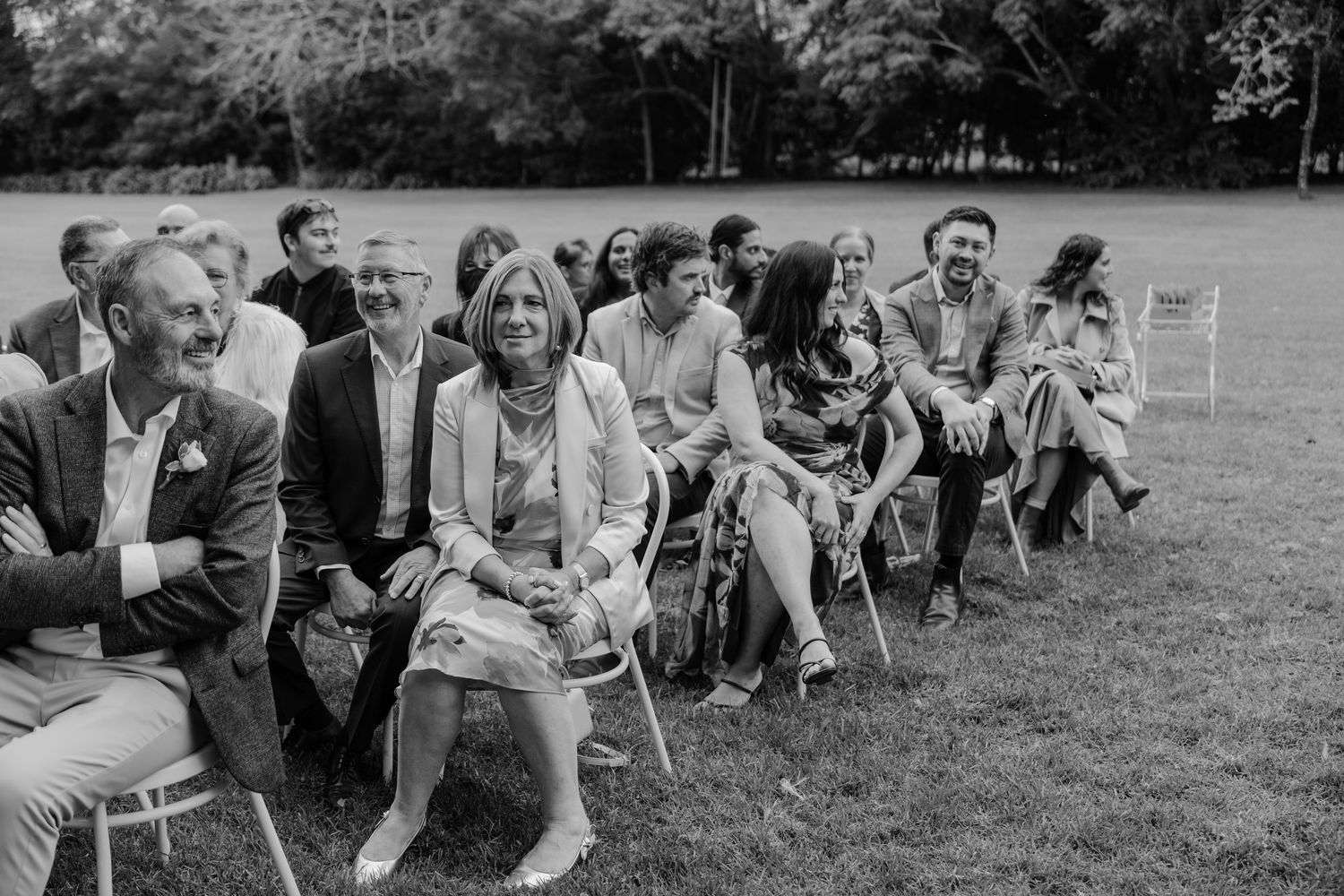 Black and white photo of wedding guests seated on chairs outdoors, smiling and watching a ceremony on a green lawn.