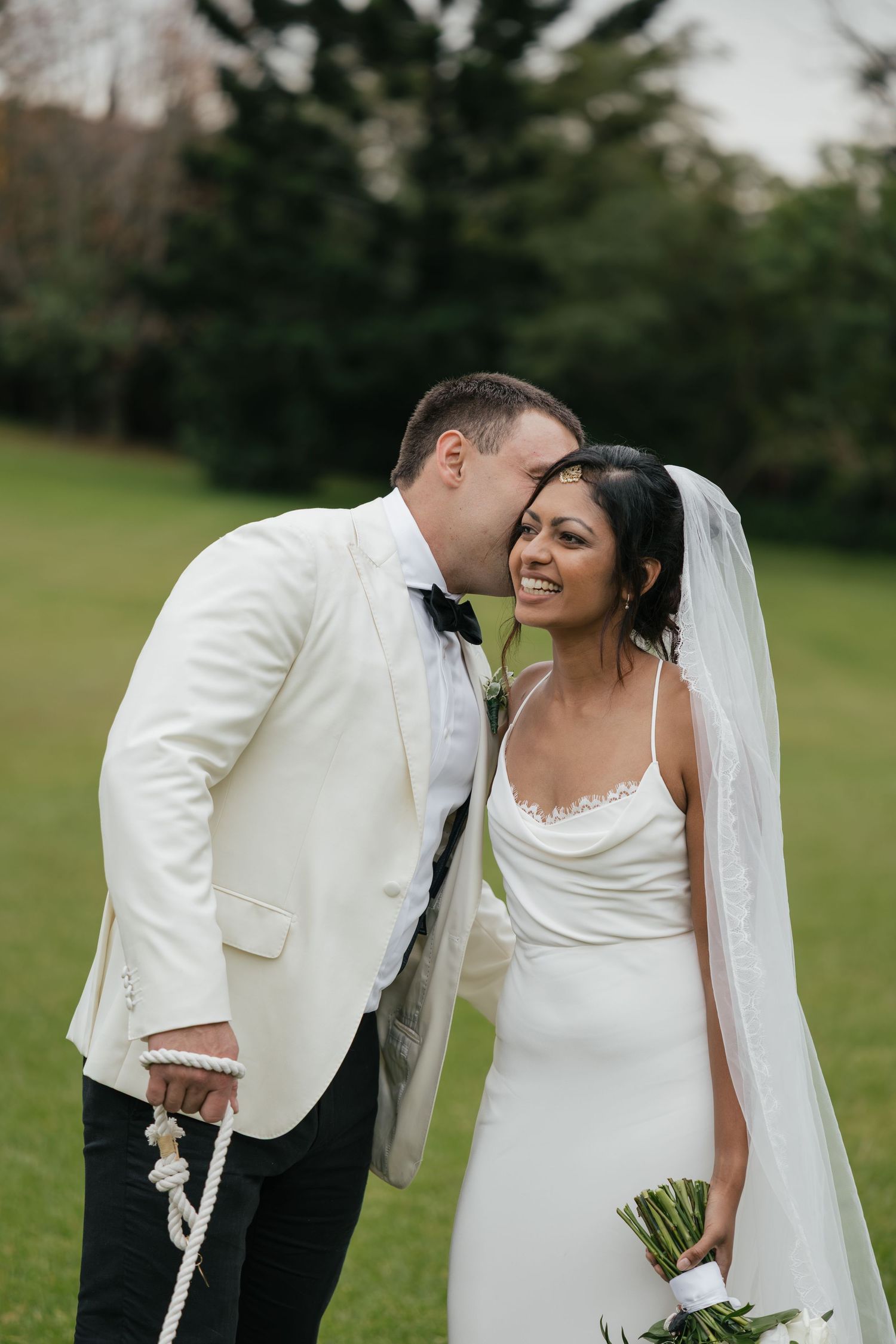 Bride in white gown and veil smiles as groom in ivory tuxedo kisses her cheek on lush green lawn.