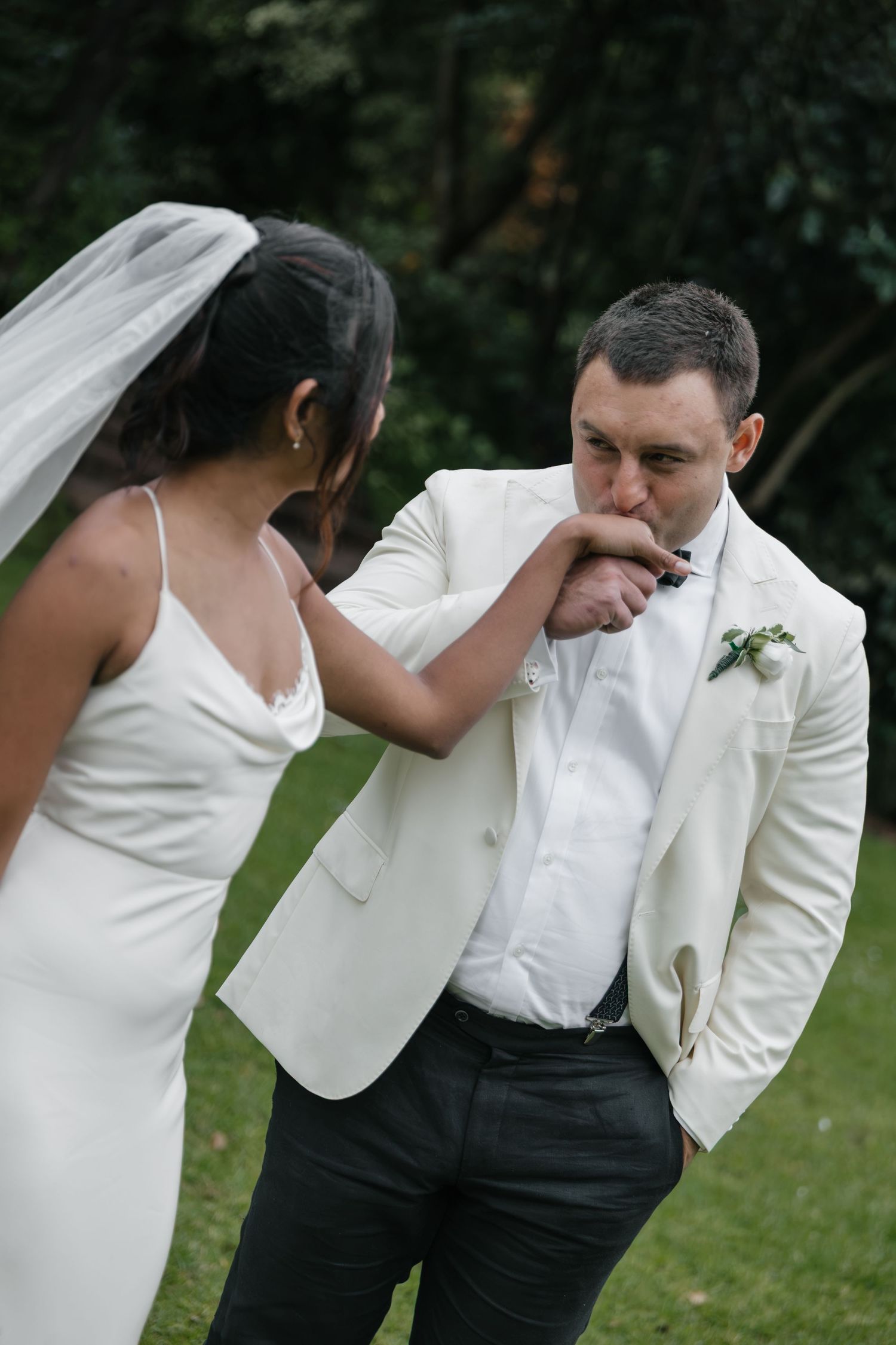 Groom in white tuxedo jacket kissing bride's hand during outdoor wedding ceremony on green lawn.