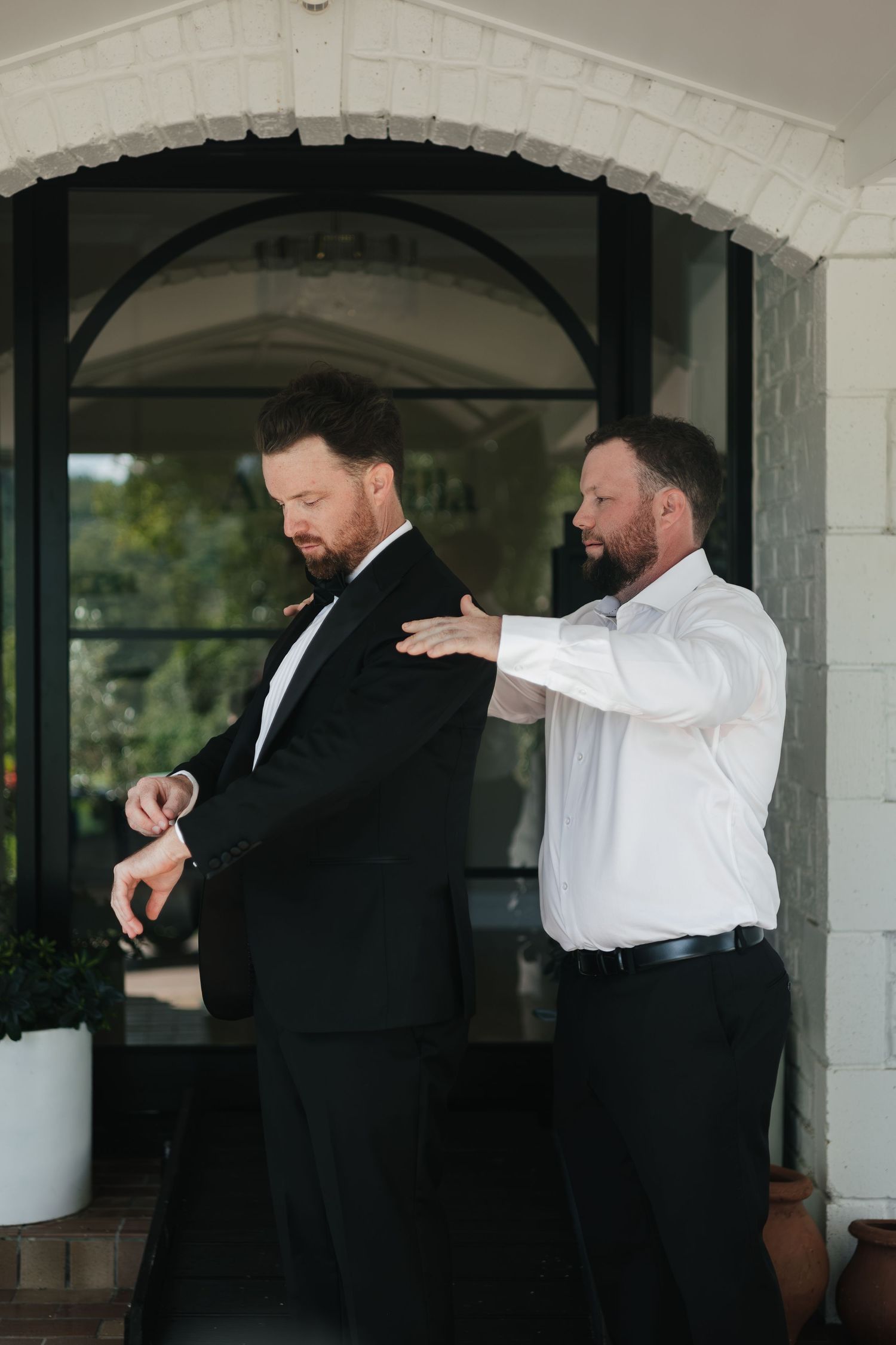 Groom in black tuxedo checks watch while groomsman in white shirt helps him prepare before wedding ceremony.