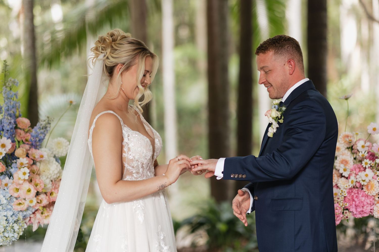 Bride placing wedding ring on groom's finger during outdoor ceremony with floral backdrop.