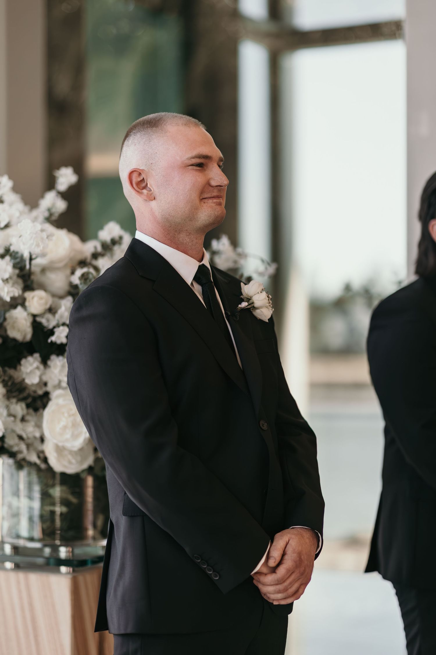 A groom in a black suit stands smiling at the altar with white floral arrangements in the background.