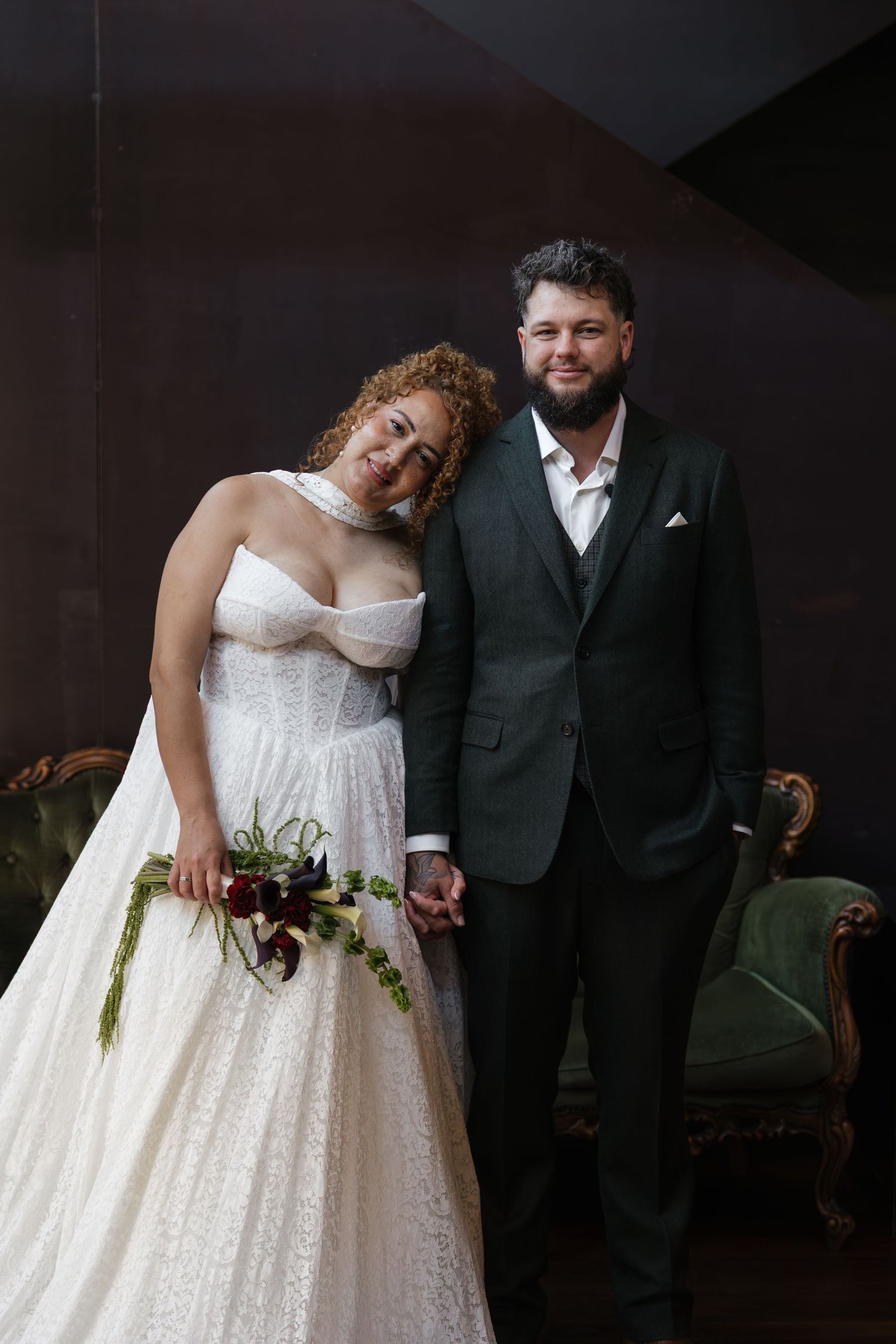 A bride in a white lace gown holding a bouquet stands beside a groom in a dark suit, posing together indoors.