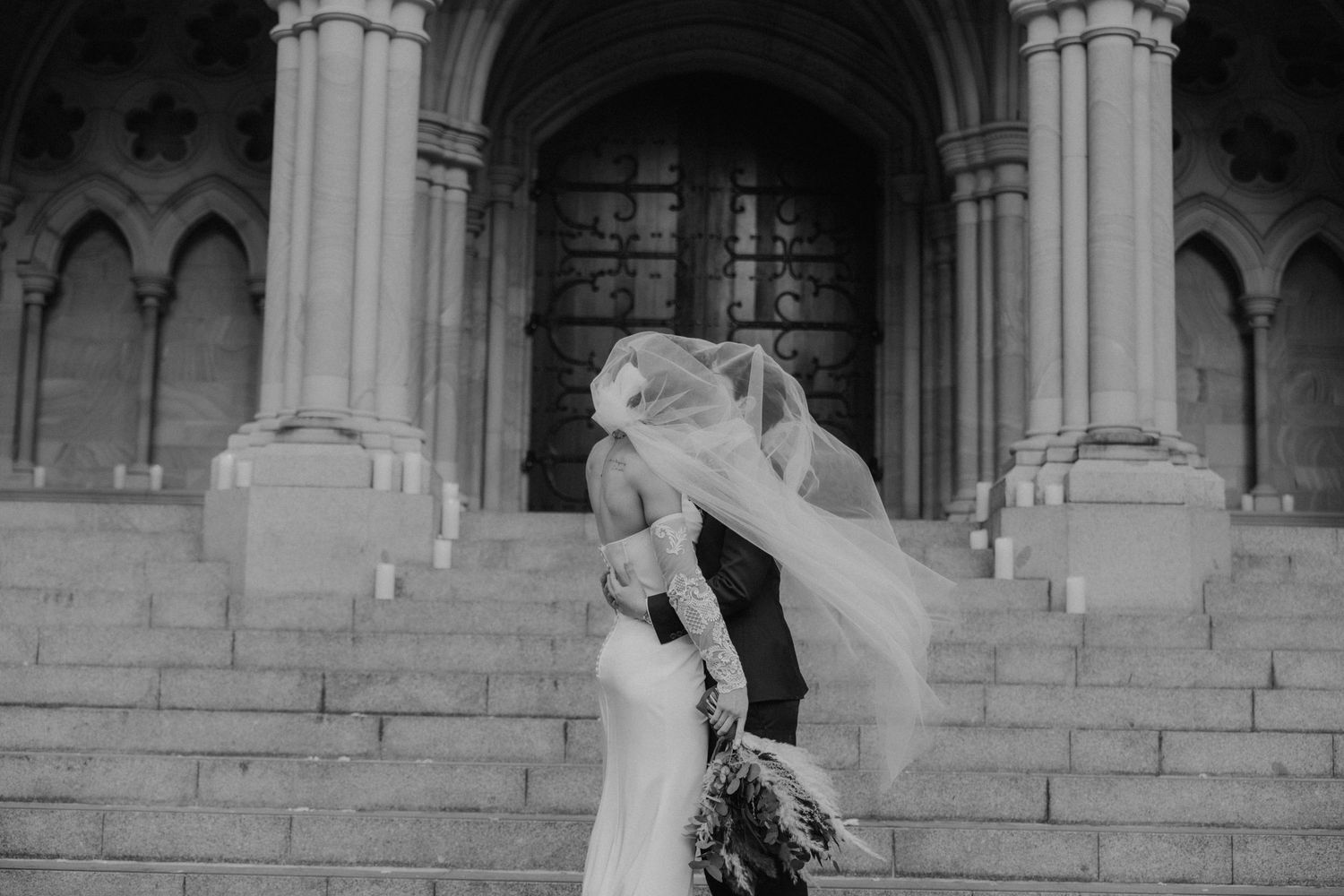 Black and white photo of bride and groom embracing on church steps, veil blowing in wind.