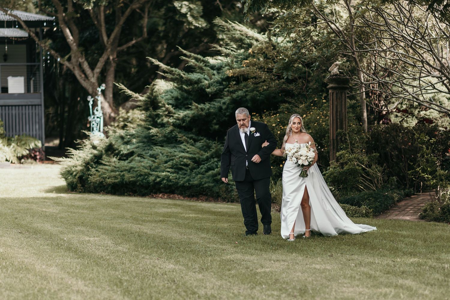 Bride in white gown walks with father across green lawn past evergreen trees at outdoor wedding ceremony.