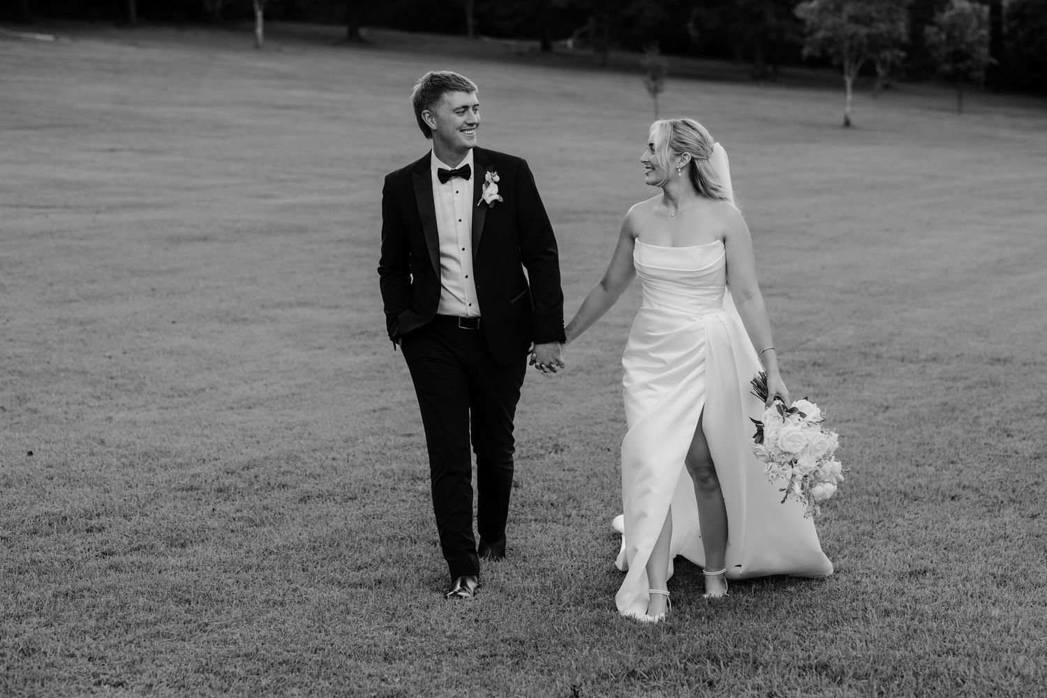 Black and white photo of a bride and groom walking hand in hand across a grassy lawn on their wedding day.