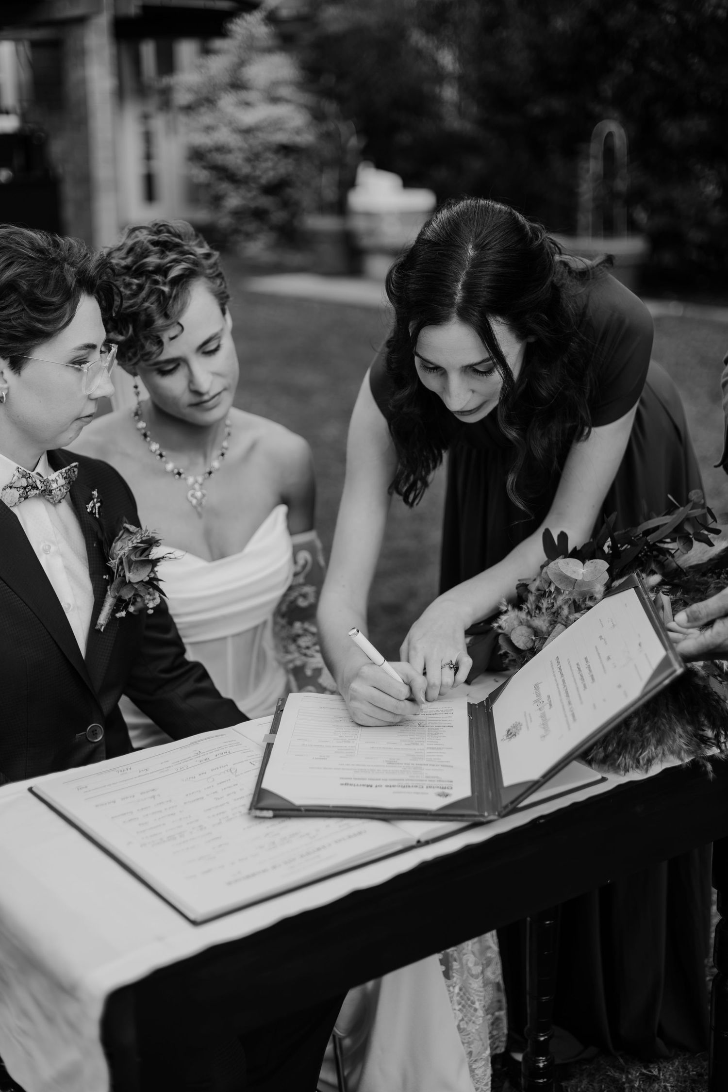 Two brides and a witness sign wedding documents at an outdoor ceremony table in black and white photo.