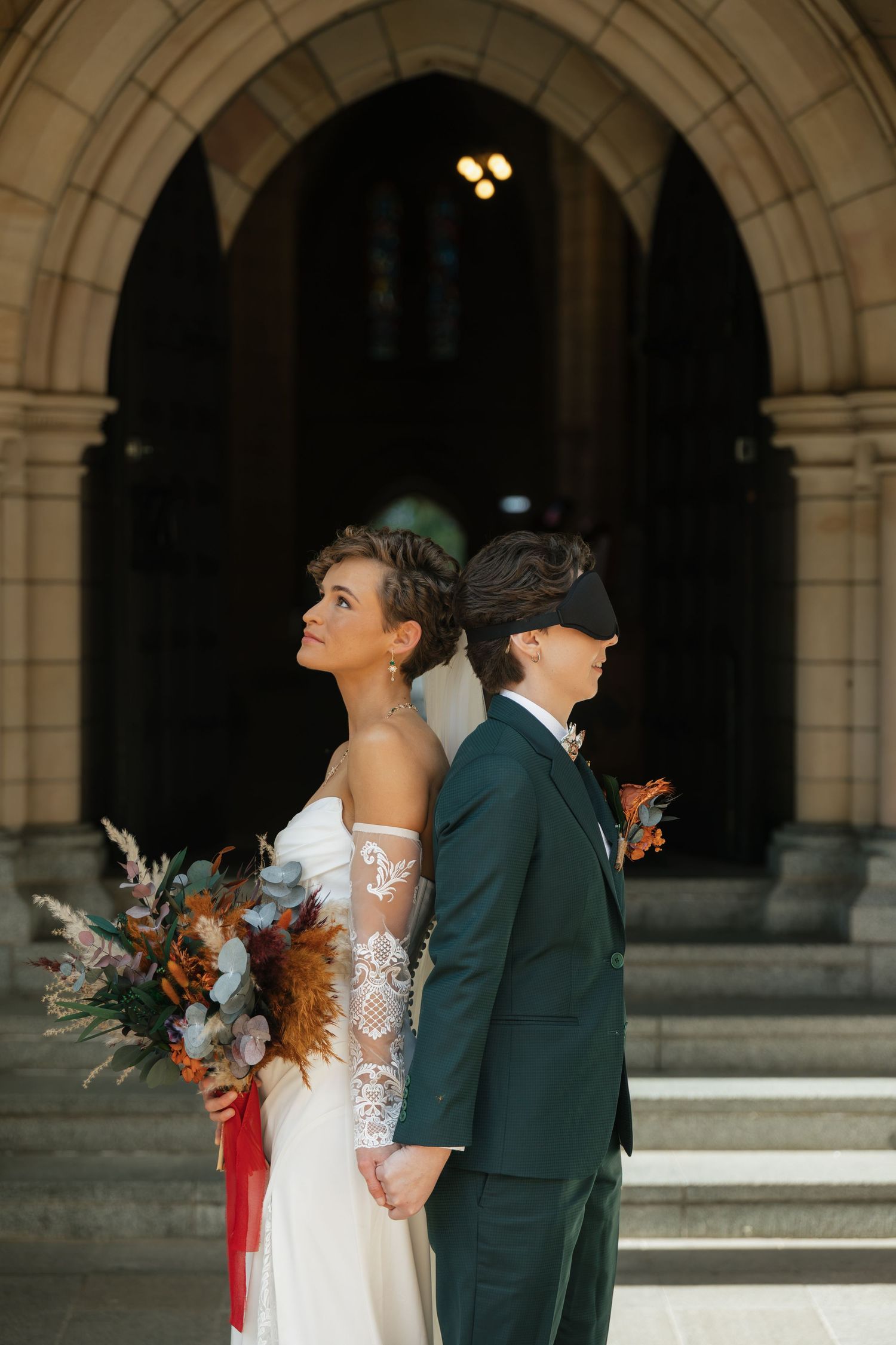 Two brides stand back-to-back on church steps, one in lace gown with bouquet, other in dark suit with blindfold.