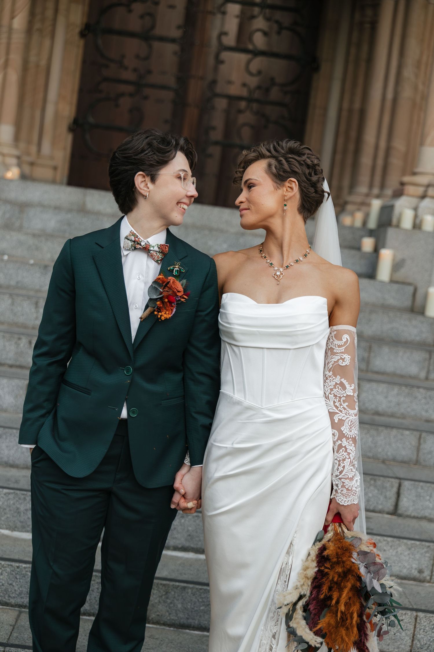 Two brides hold hands on church steps, one in a green suit with boutonniere, the other in a white strapless gown with lace sleeve.