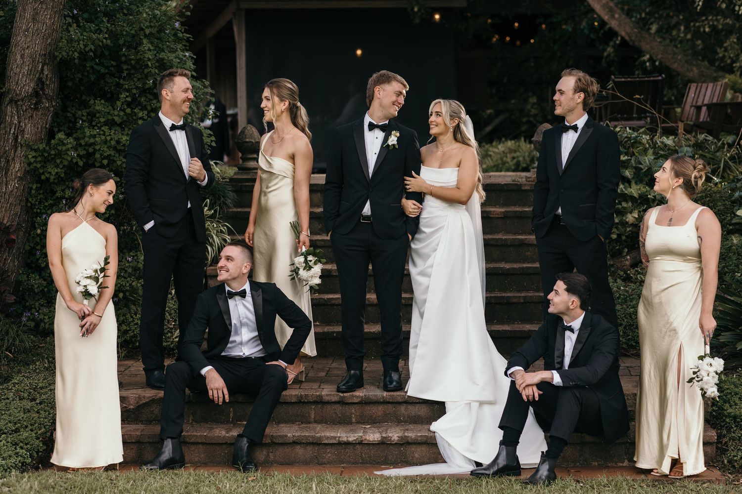 Wedding party poses on stone steps, bride and groom center in white gown and tuxedo, bridesmaids in gold dresses, groomsmen in black suits.