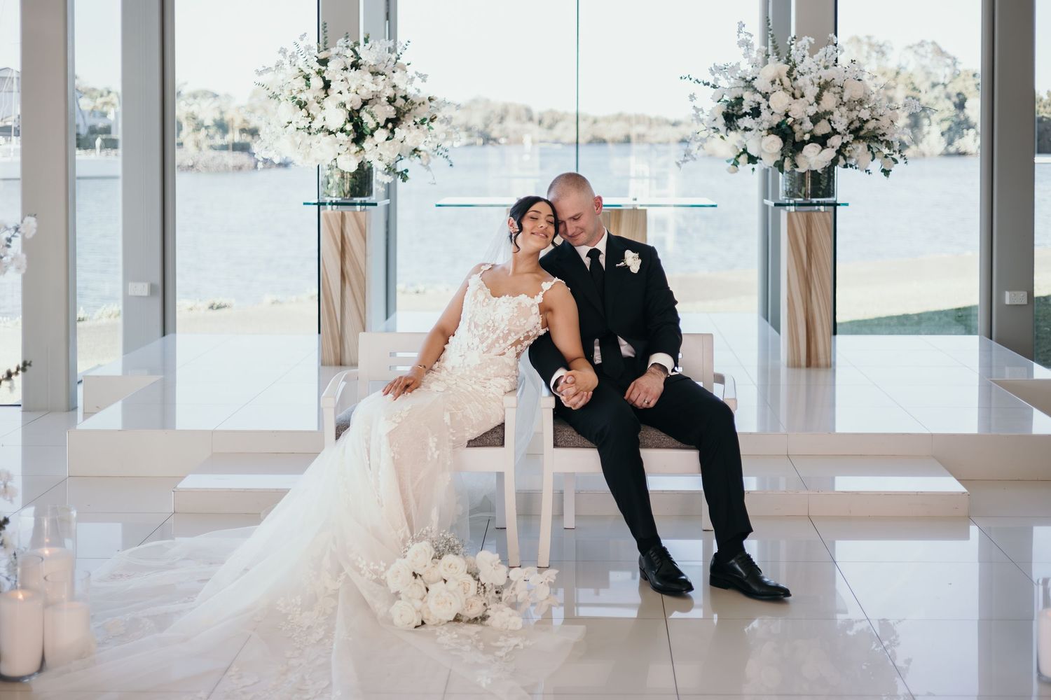 Bride and groom sit together on white chairs at elegant waterfront wedding ceremony with white floral arrangements.