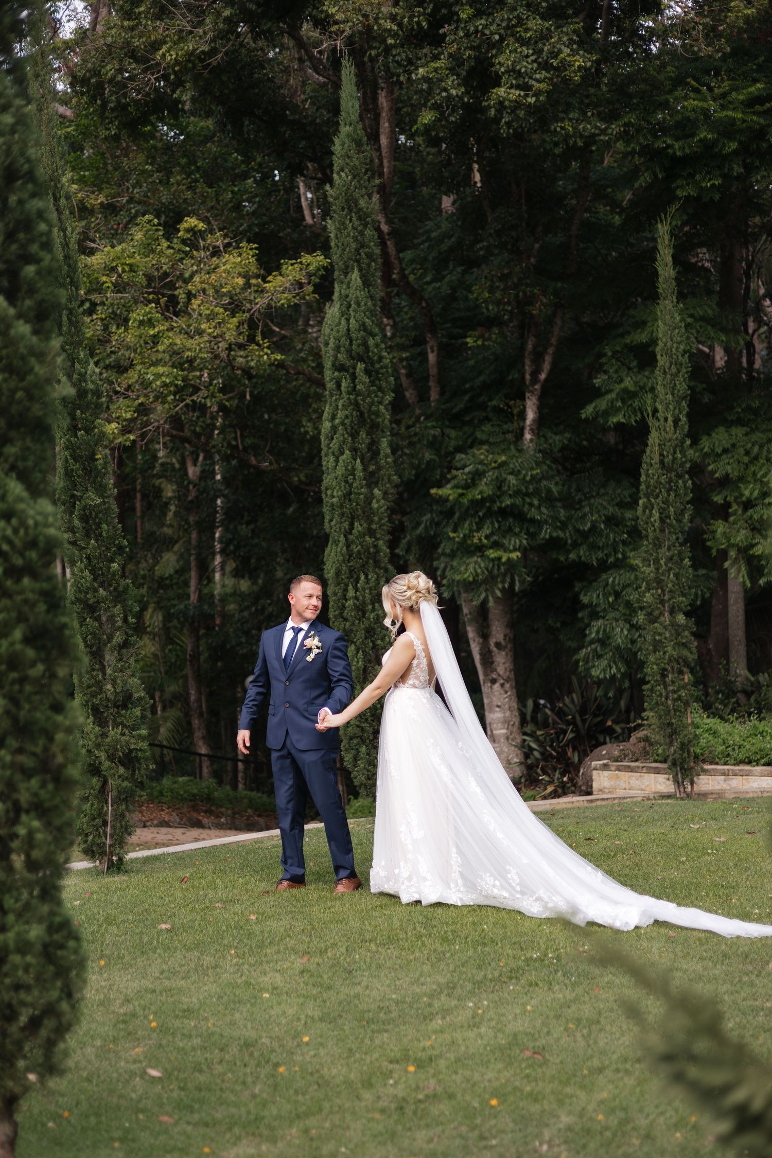 Bride and groom holding hands in lush garden with tall cypress trees, bride wearing flowing white gown with long veil.