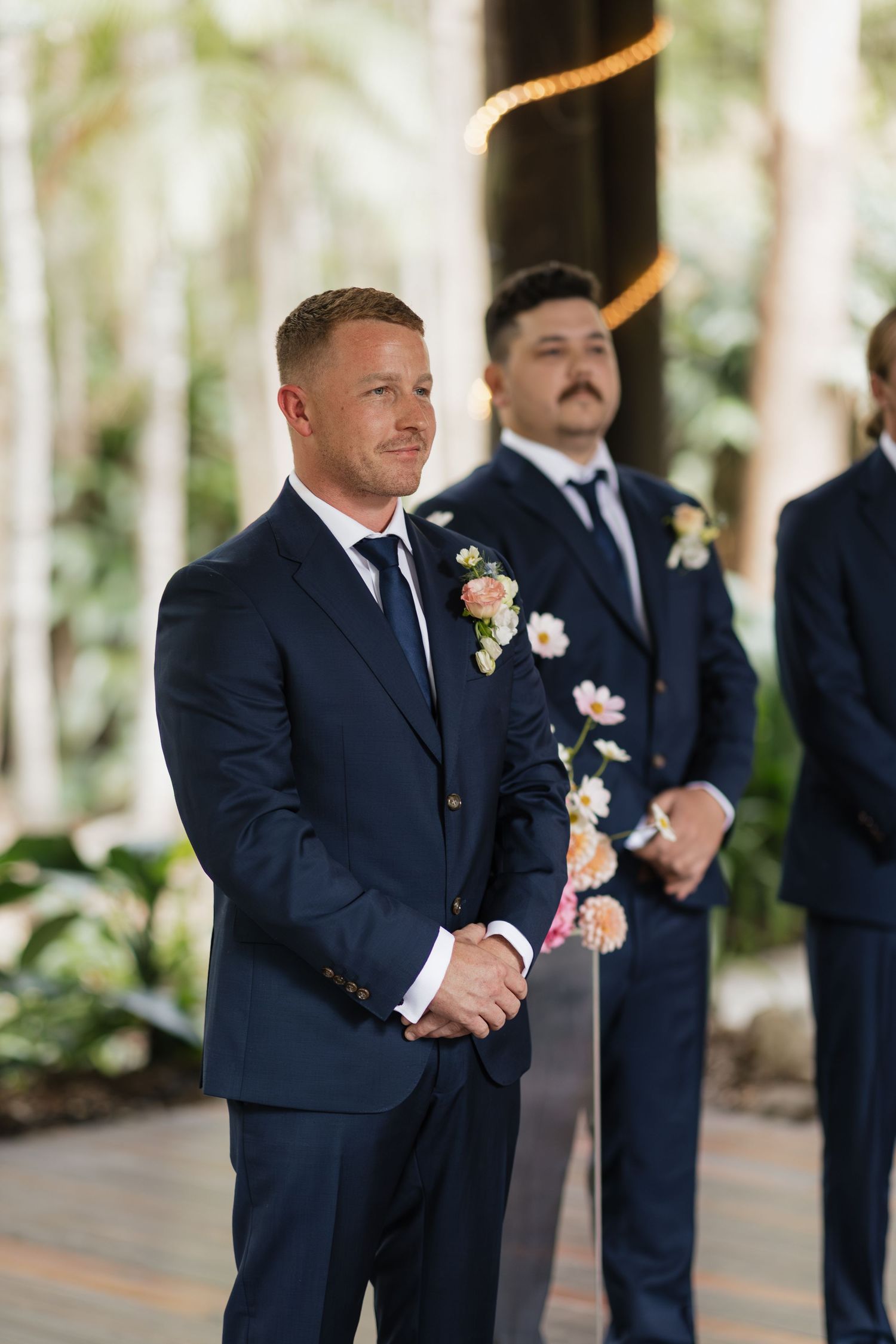 Groom in navy suit smiling emotionally at altar with groomsmen holding floral boutonnieres at outdoor wedding ceremony.