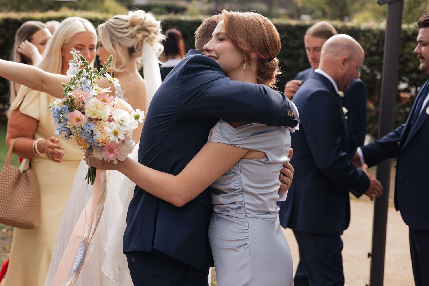 Bride and groom share a warm hug with a guest at their outdoor wedding reception, surrounded by elegantly dressed attendees.