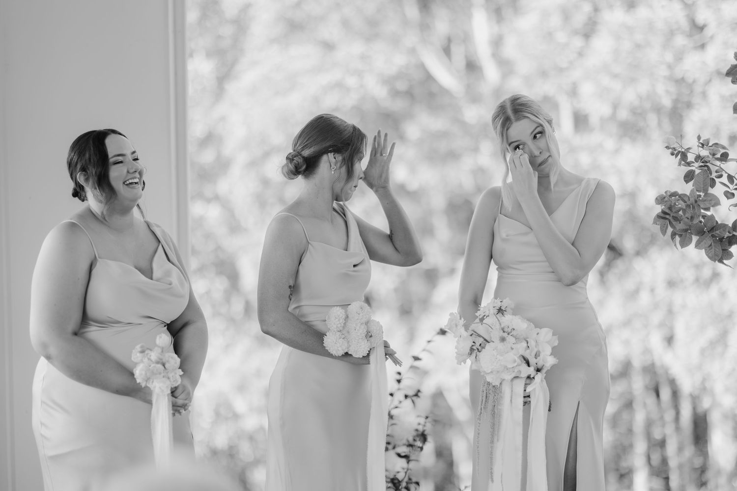 Three bridesmaids in elegant dresses laughing and crying with joy at outdoor wedding ceremony.