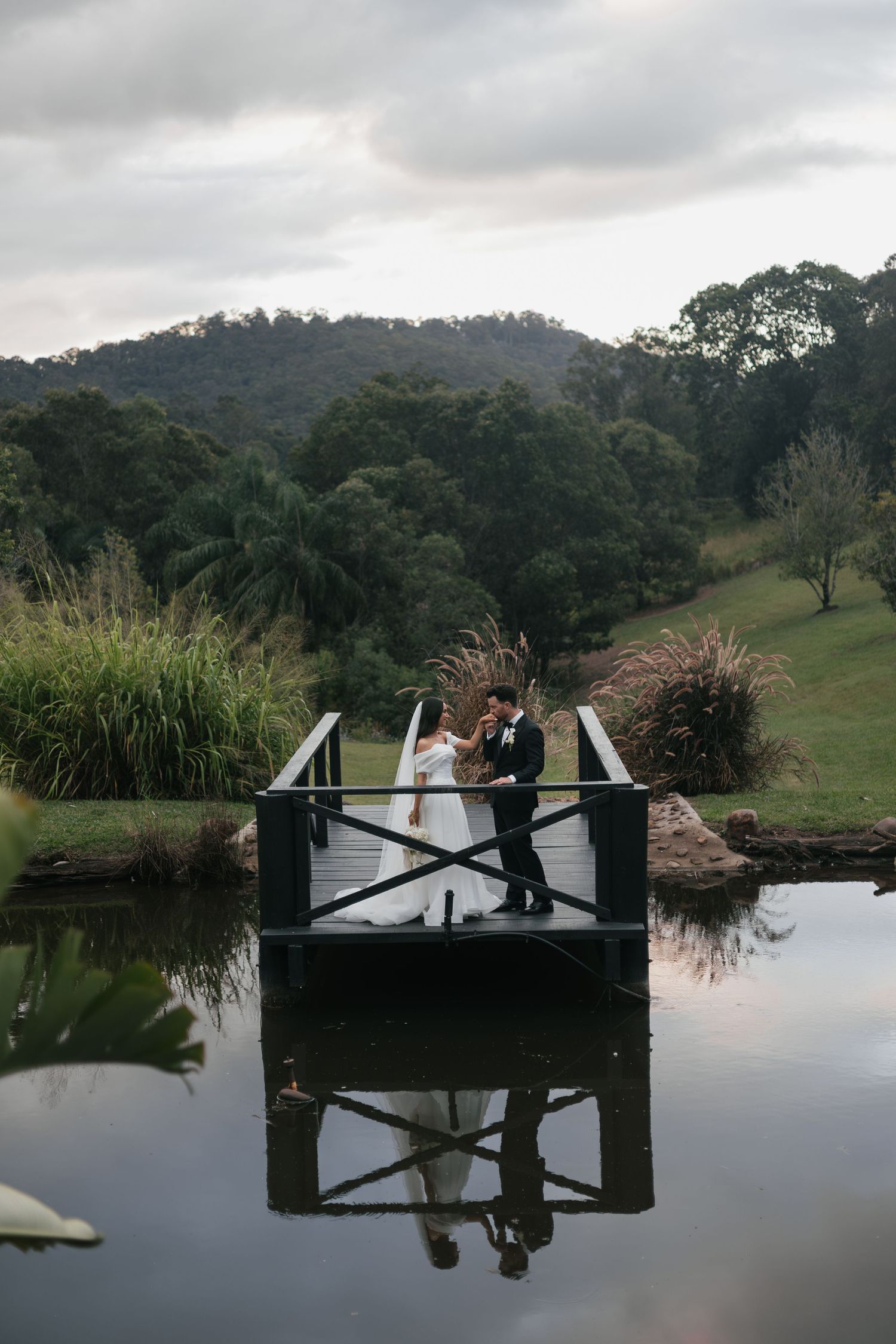 Bride and groom stand on a rustic bridge over a reflective pond, surrounded by lush green hills on their wedding day.