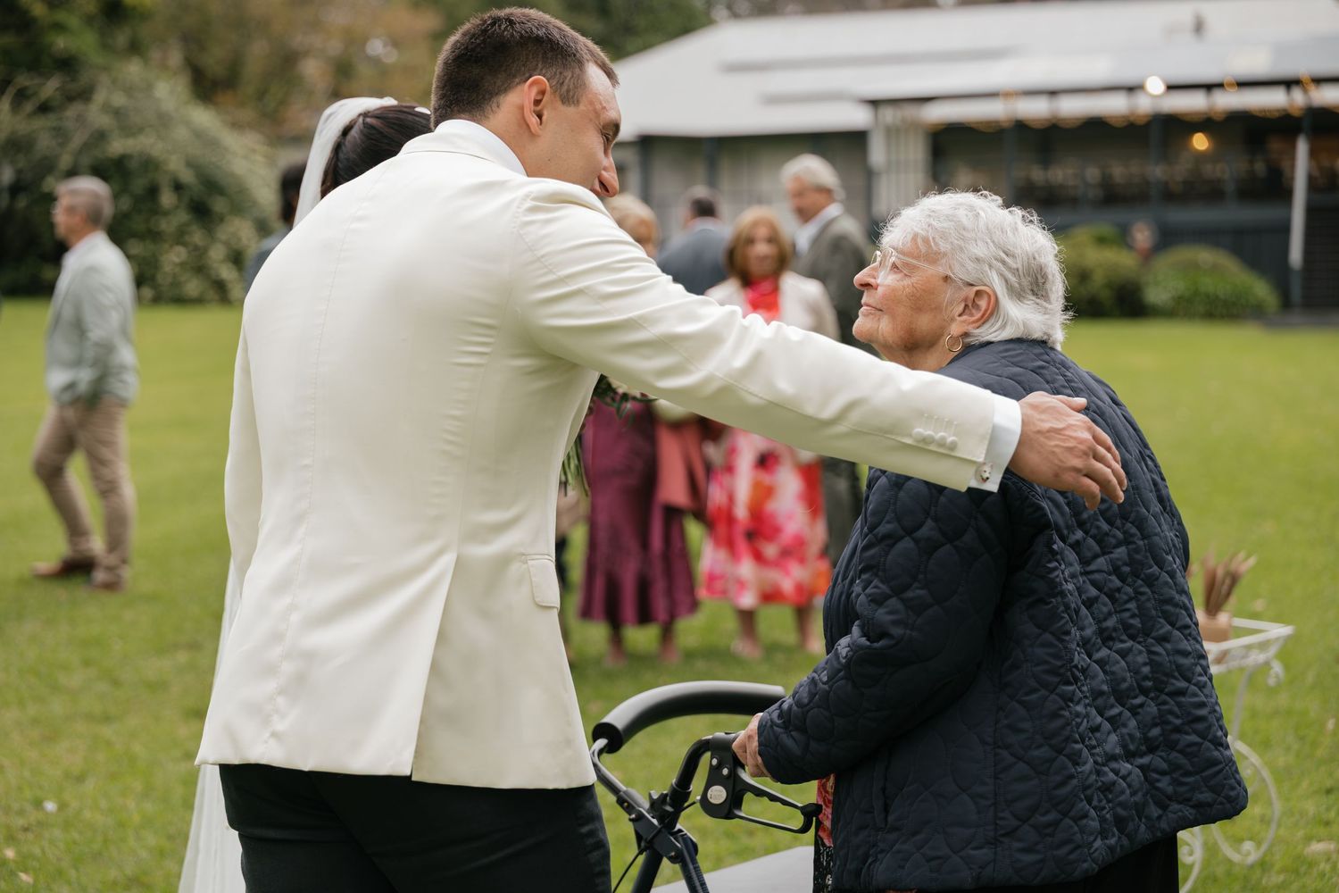 Groom in white jacket embracing elderly woman at outdoor wedding reception on green lawn.