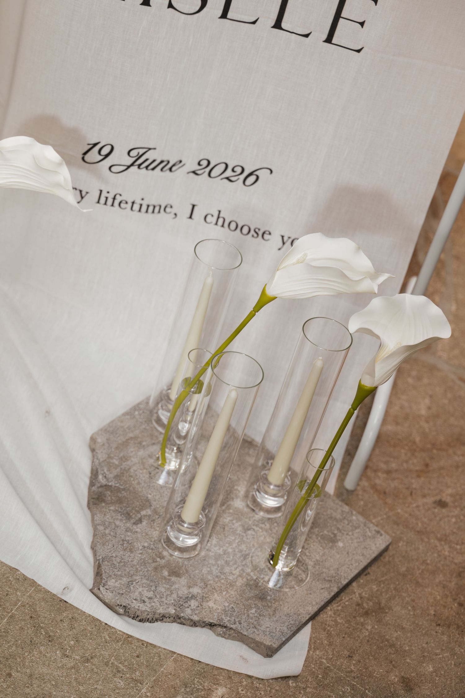 Elegant wedding decor with white calla lilies in glass vases on stone, beside a sign reading 19 June 2026.